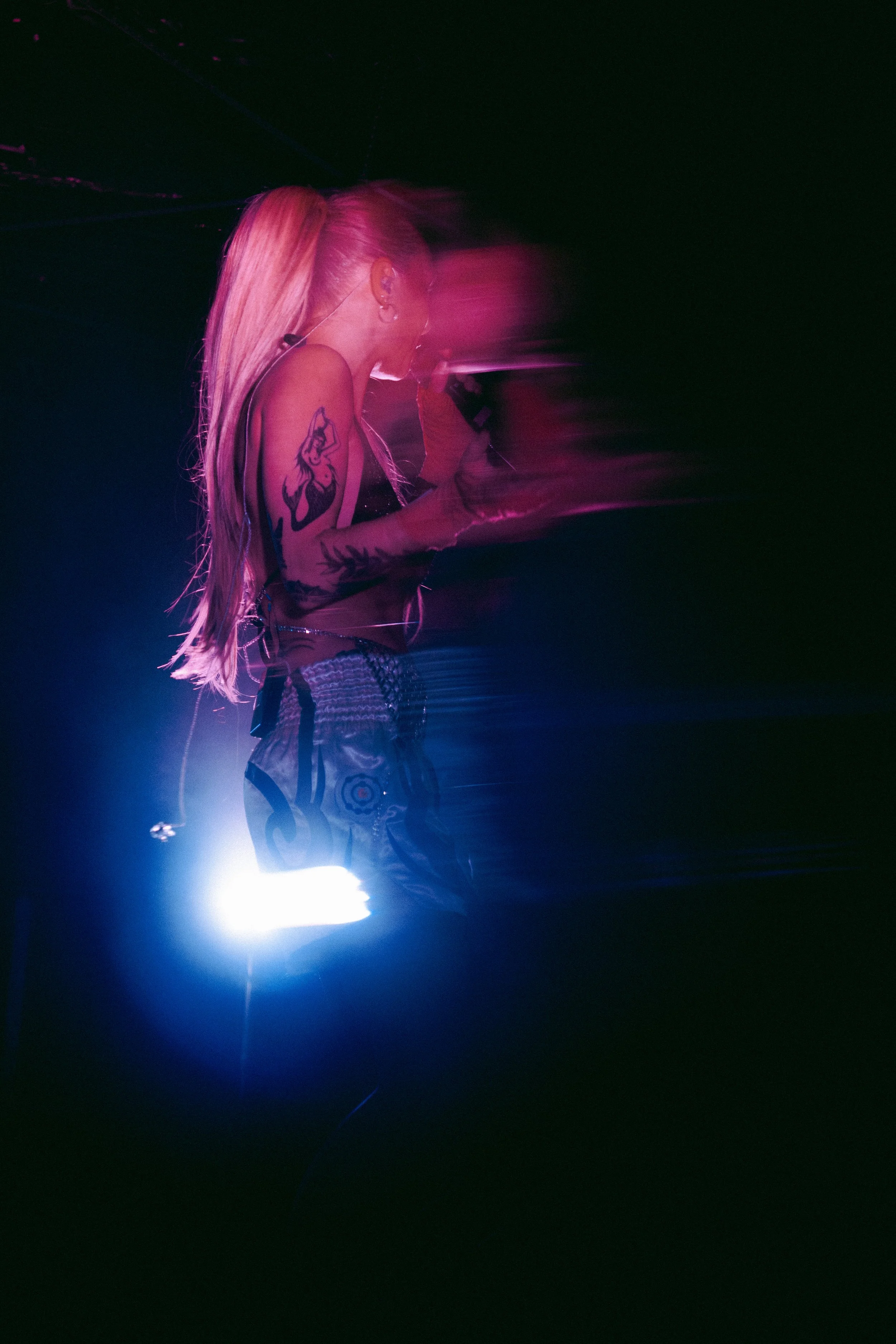A woman with long blonde hair and tattoos on her arm, wearing a black top and patterned shorts, holds a microphone on stage with pink and purple lights creating a blurred motion effect.