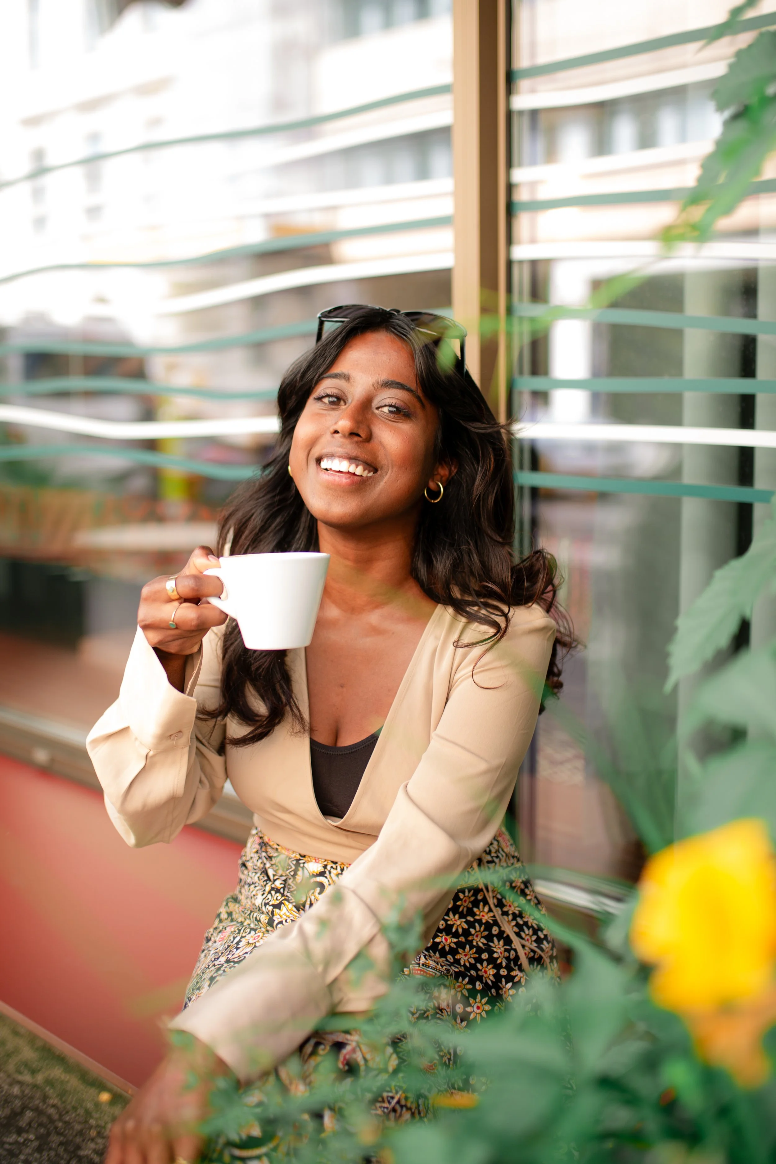 A woman with dark hair and hoop earrings, smiling and holding a white coffee mug, sitting near a window with horizontal blinds, surrounded by greenery and yellow flowers.