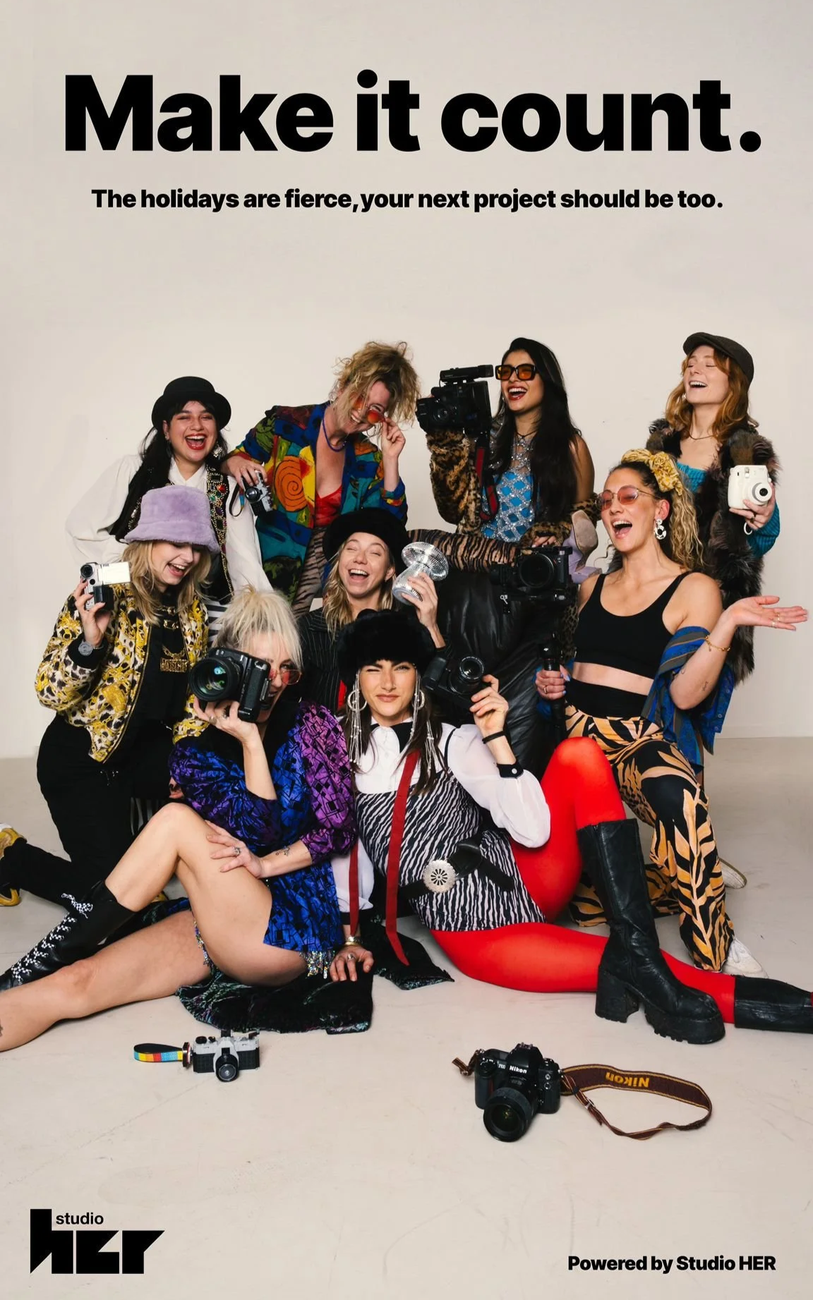 Group of women dressed in colorful, eclectic outfits posing with cameras and laughing together in a studio with a plain background.