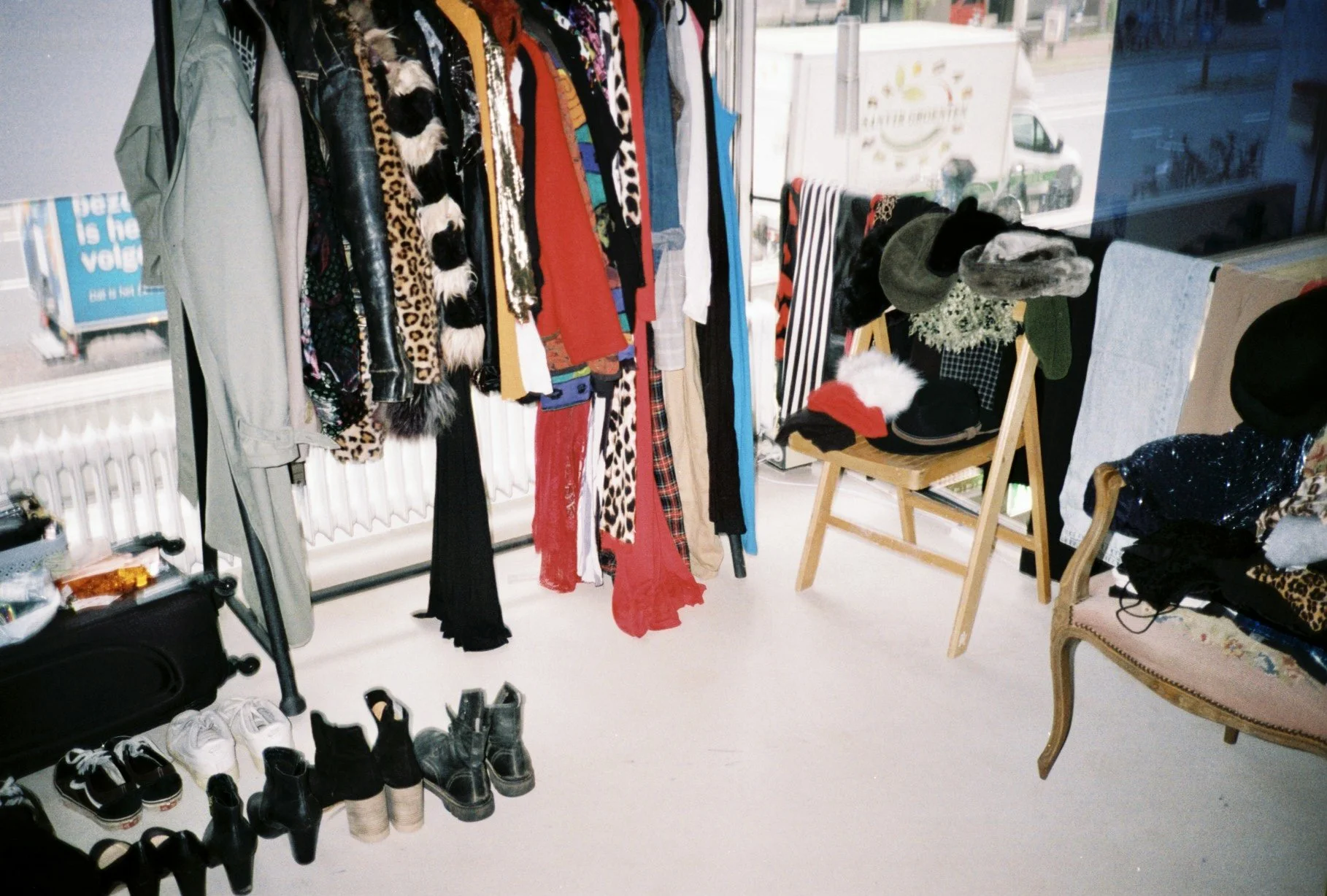 Clothes hanging on racks and a chair with various hats inside a store window display.