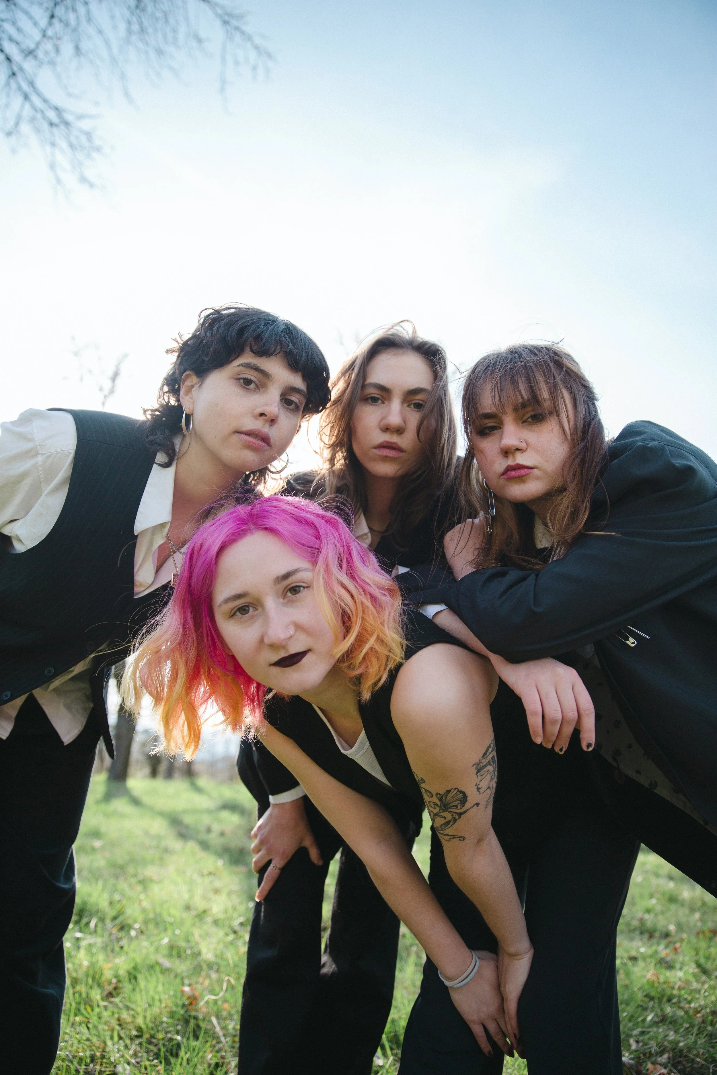 Four women with different hair colors and styles posing outdoors in a grassy field on a clear day.