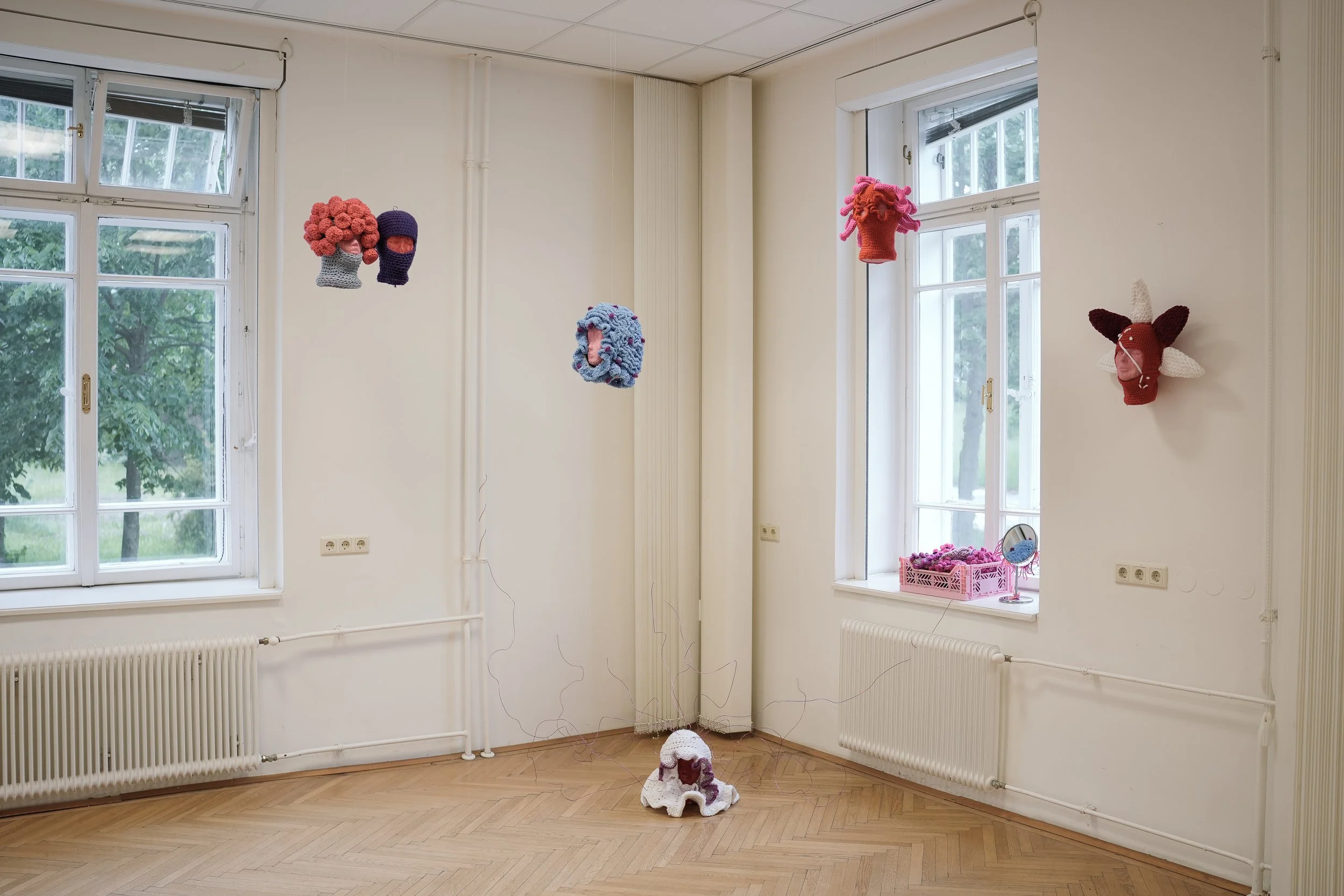 An art installation in a room with white walls and wooden floor, featuring knitted and crocheted sculptural pieces hanging from the ceiling and mounted on the wall, including colorful abstract forms and a unicorn head.