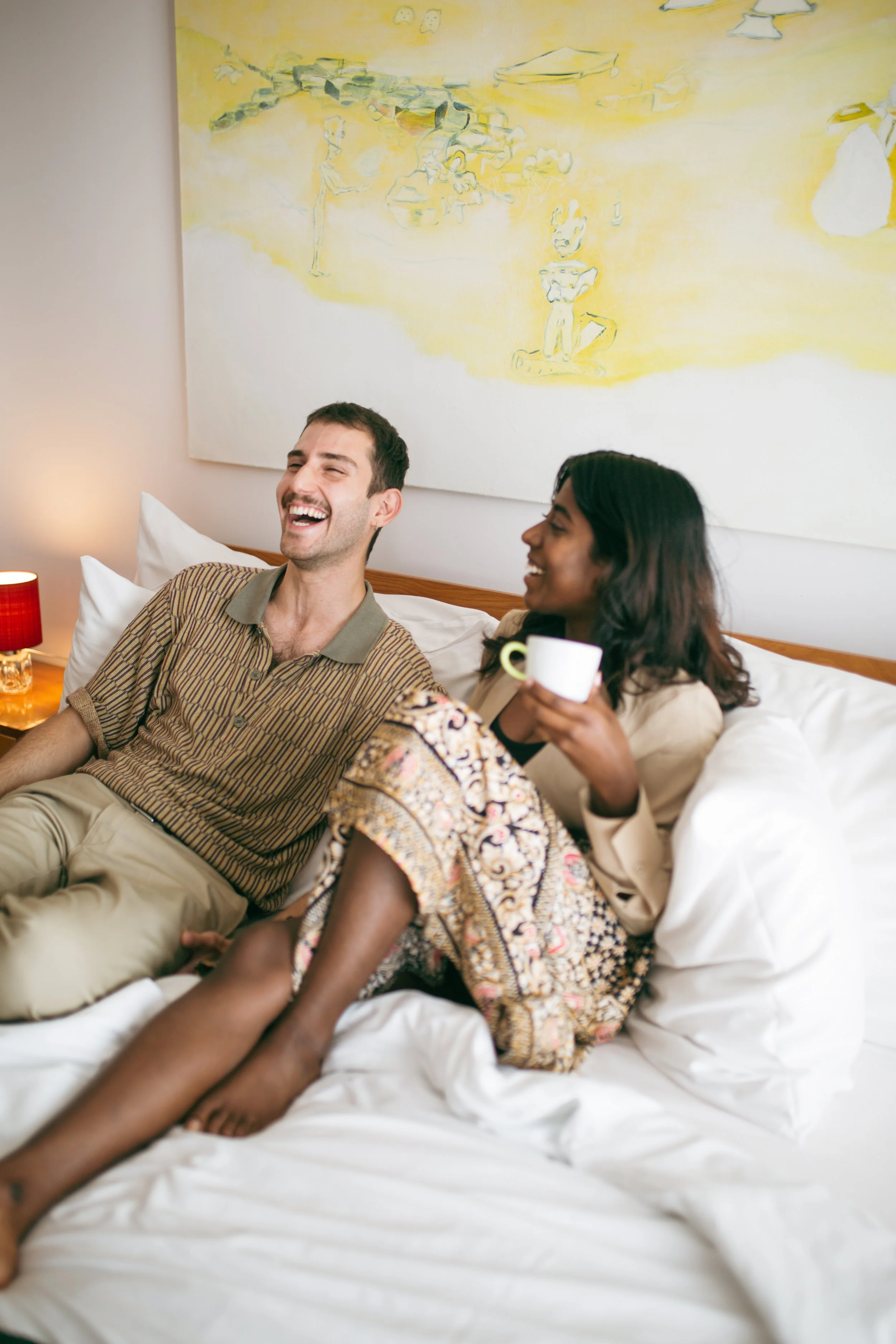 A man and woman sitting on a bed, smiling and laughing, with the woman holding a cup. There is a lamp with a red shade on a nightstand beside the bed, and abstract artwork on the wall behind them.