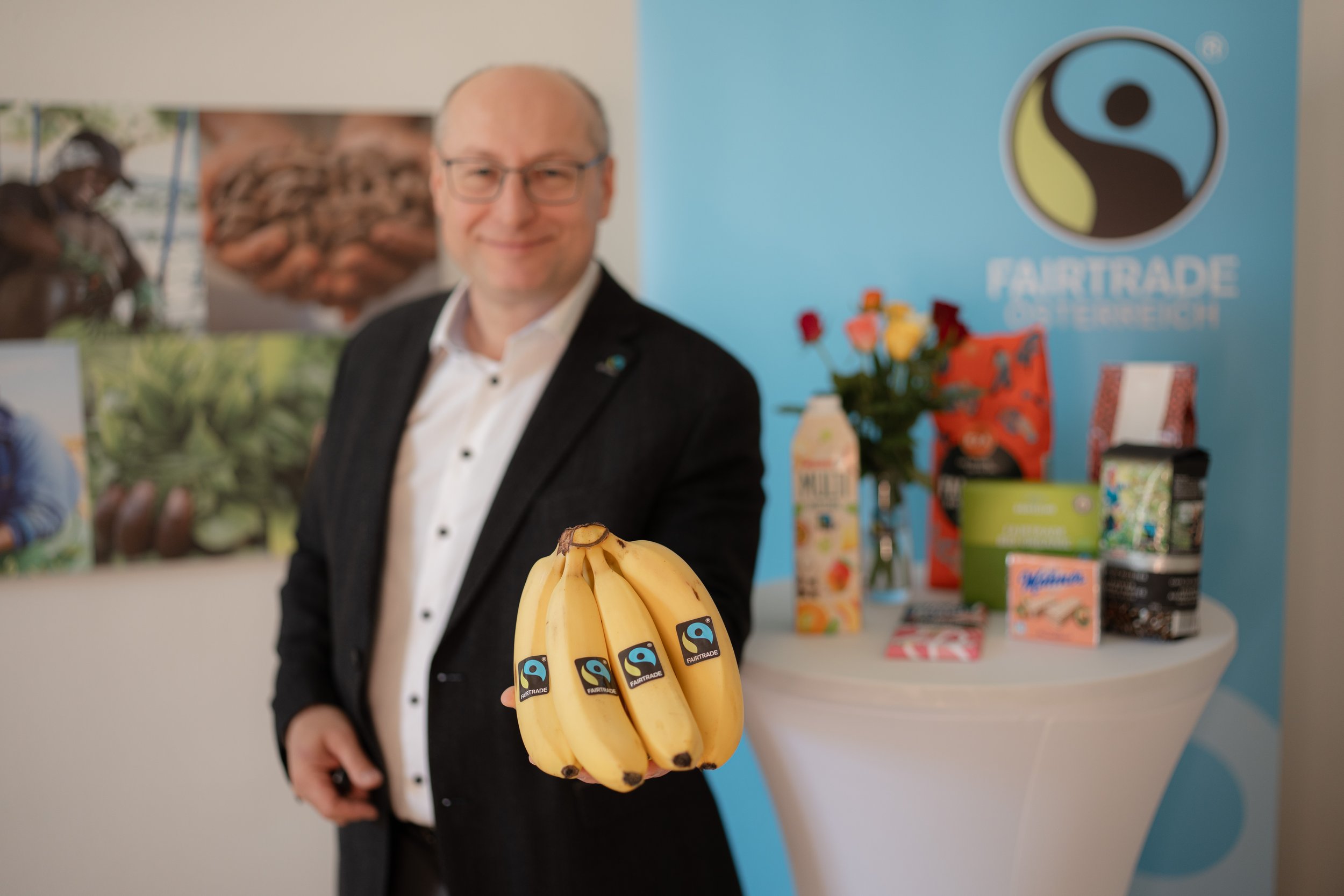 A man in a black suit holding a bunch of bananas with a Fairtrade sticker, standing in front of a display table with various products and a Fairtrade logo.