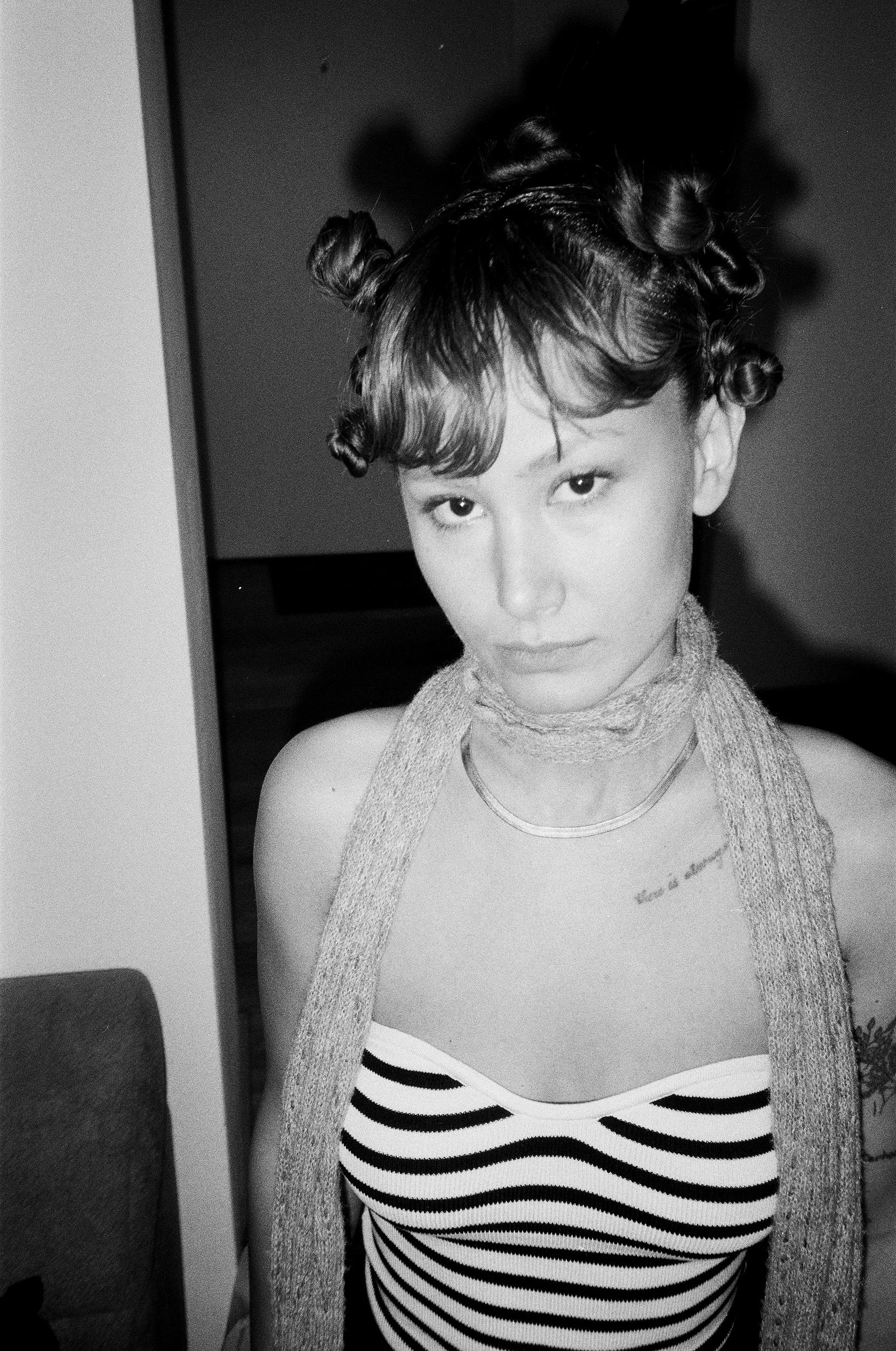 A young woman with short hair styled with multiple small buns, wearing a striped top and a scarf around her neck, looking directly at the camera.