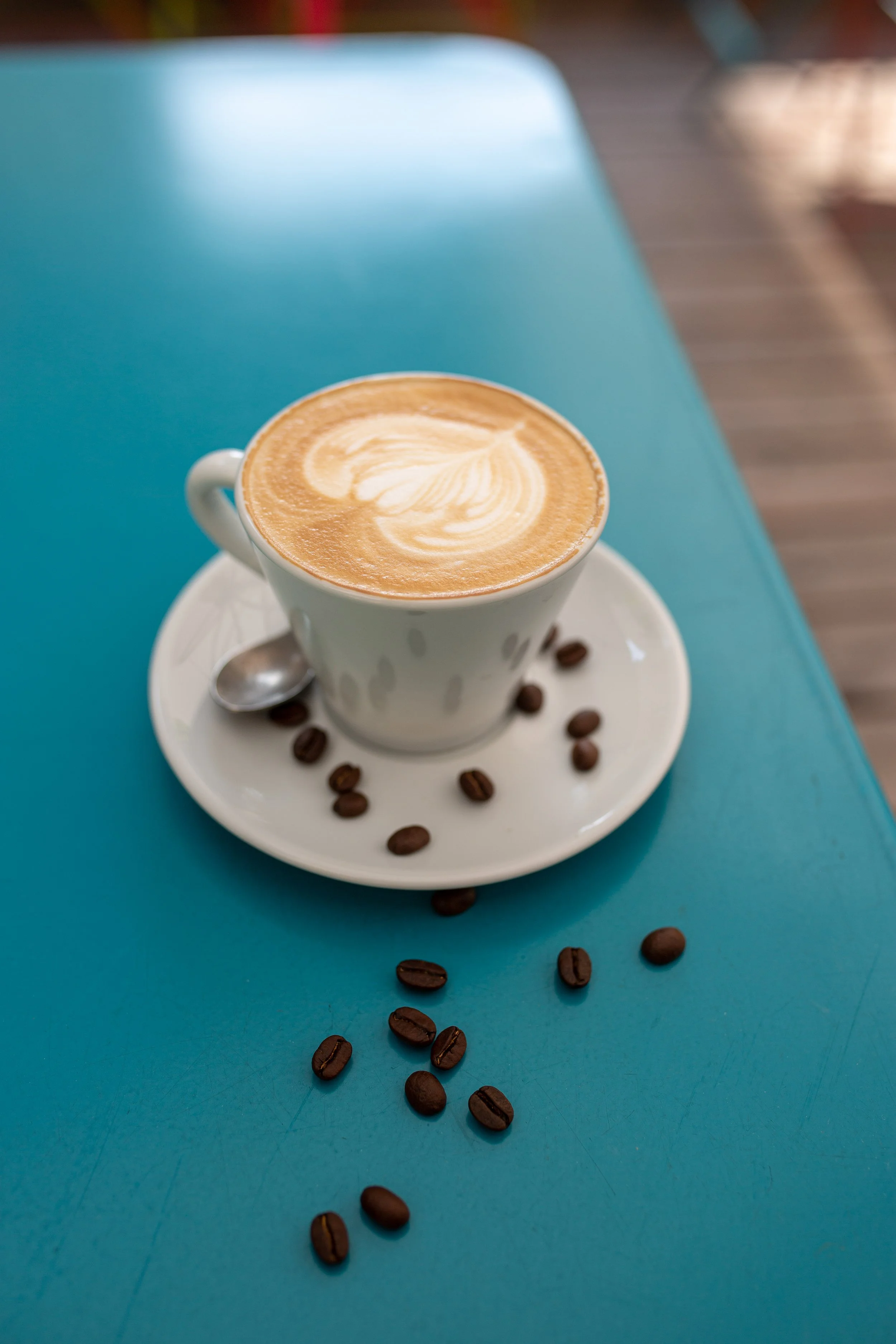 A cup of coffee with latte art, on a saucer with scattered coffee beans and a small spoon, on a turquoise table.