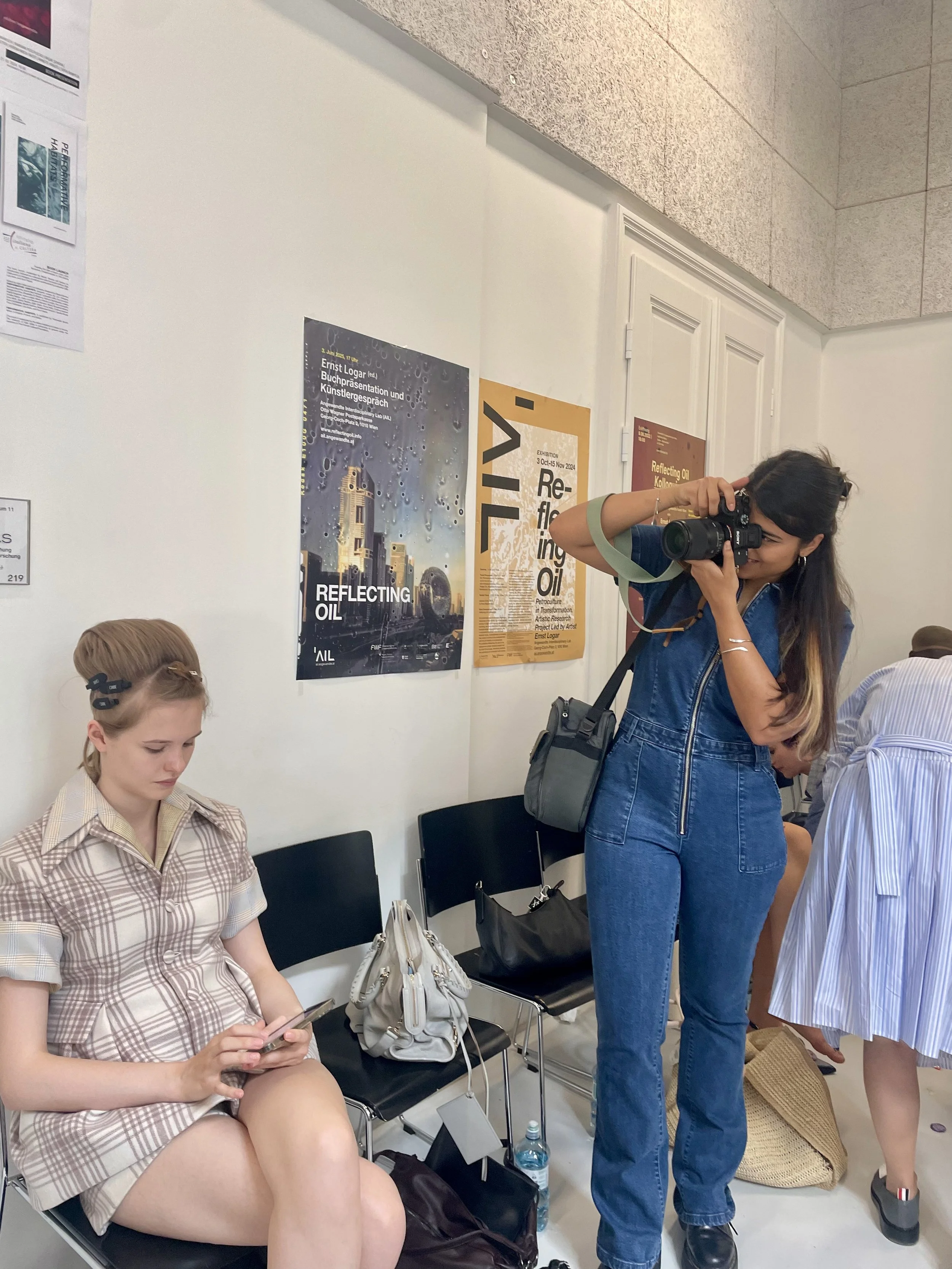 A woman taking a photo with a camera while standing next to a seated girl who is looking at her phone in a waiting area with posters on the wall.