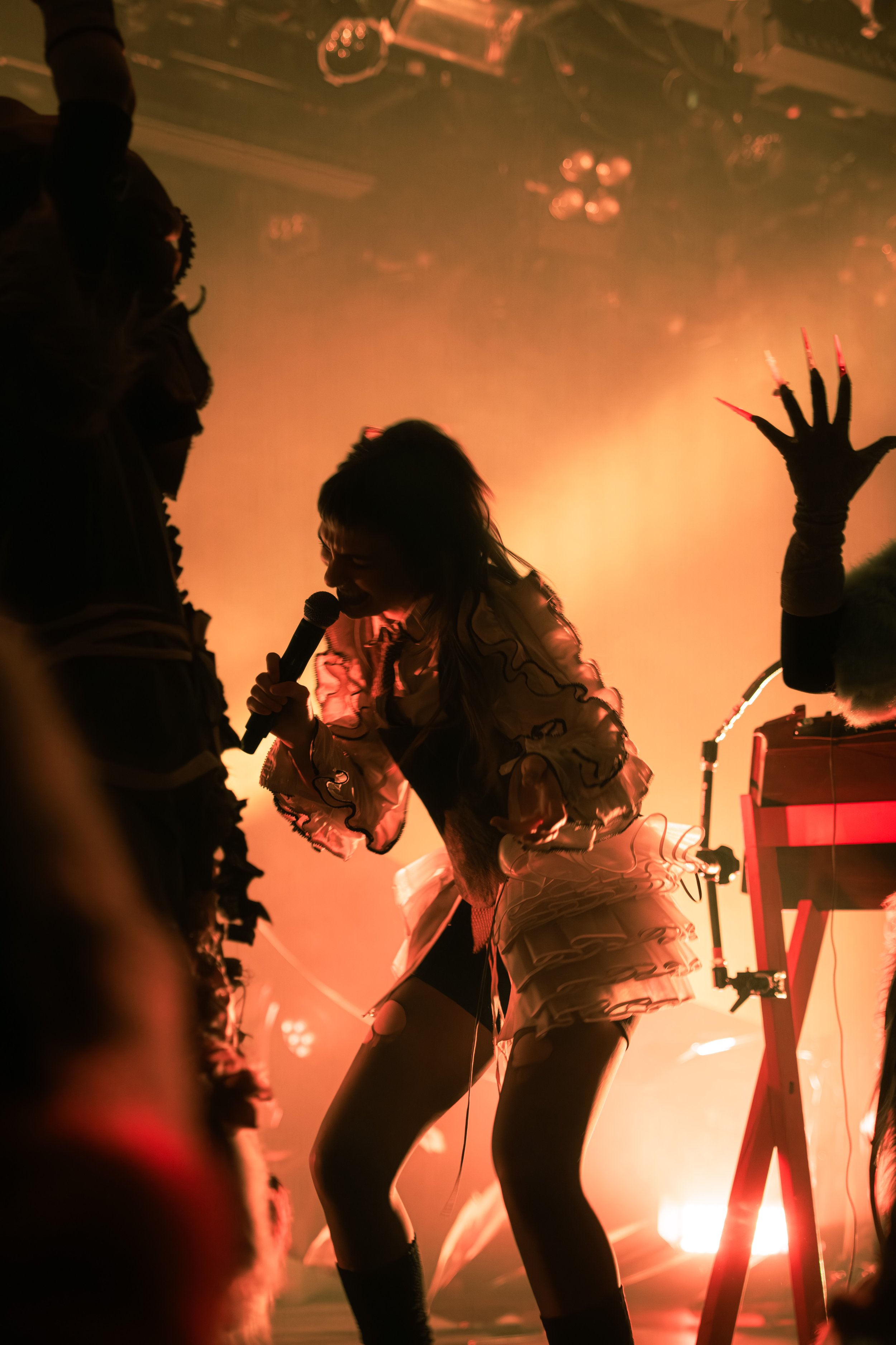 Performer singing into a microphone on stage with bright orange and yellow lighting, wearing a ruffled dress and fishnet stockings, with animated red nails.