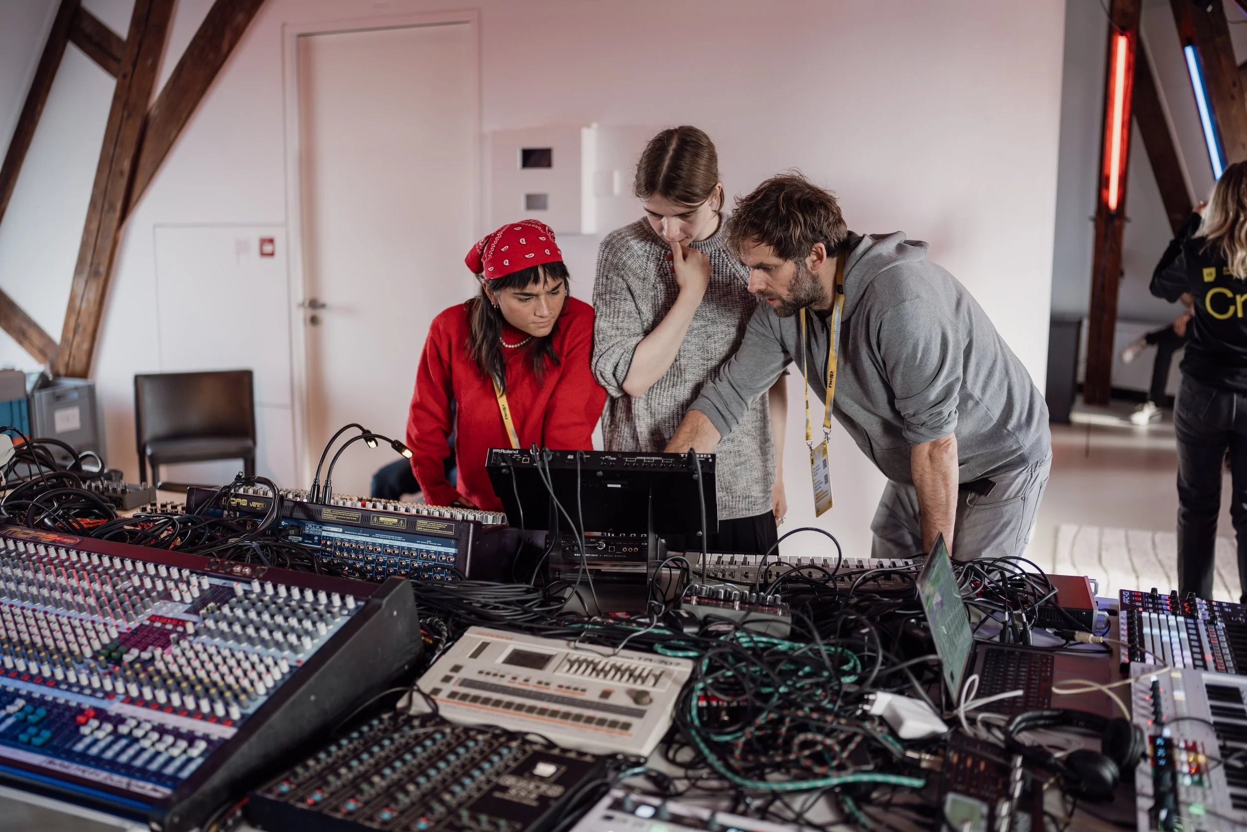 Three people working together at a table filled with audio equipment and wires, in a room with wooden beams and a white wall.