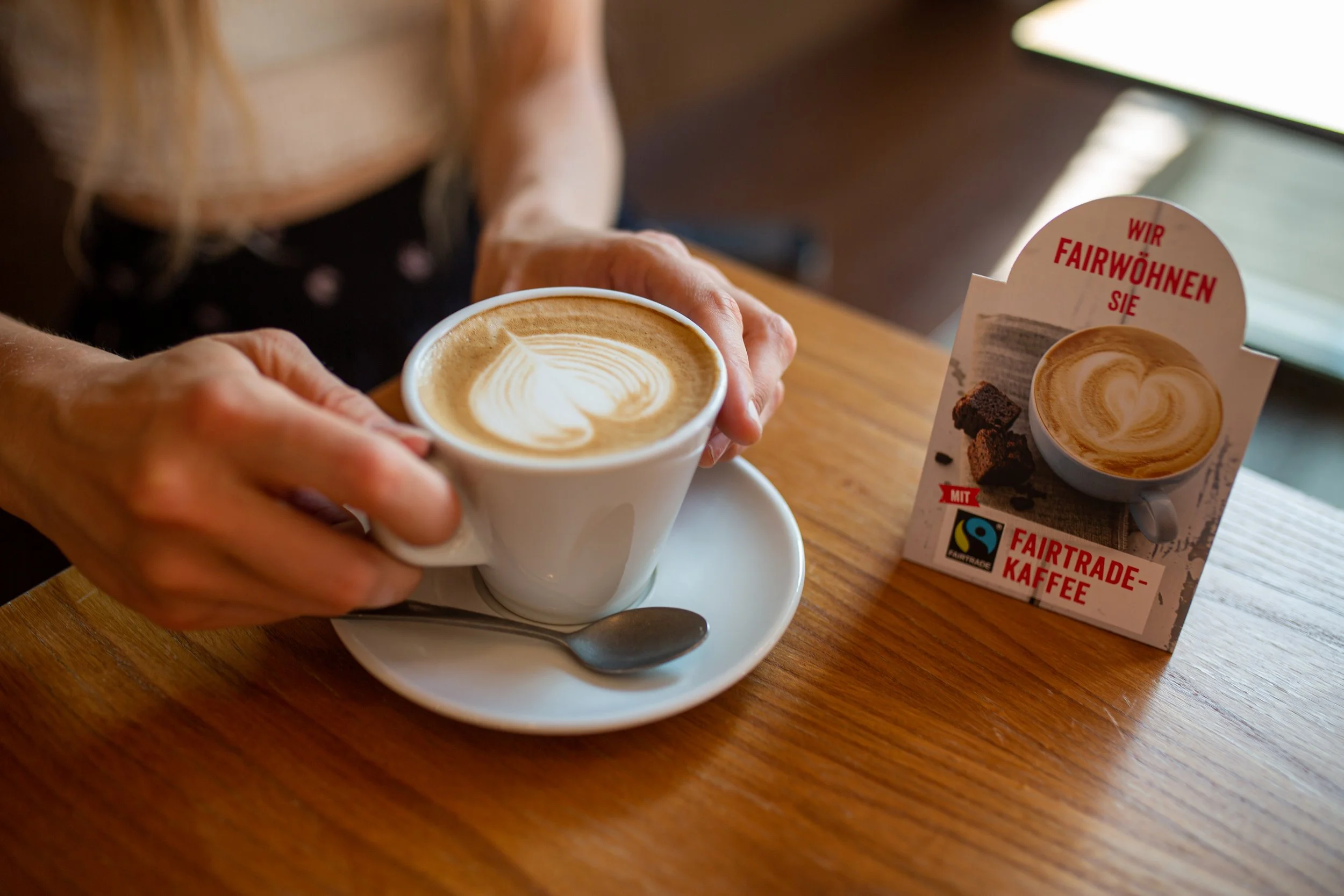 A person holding a cup of coffee with latte art, sitting at a wooden table next to a small sign promoting Fairtrade coffee.