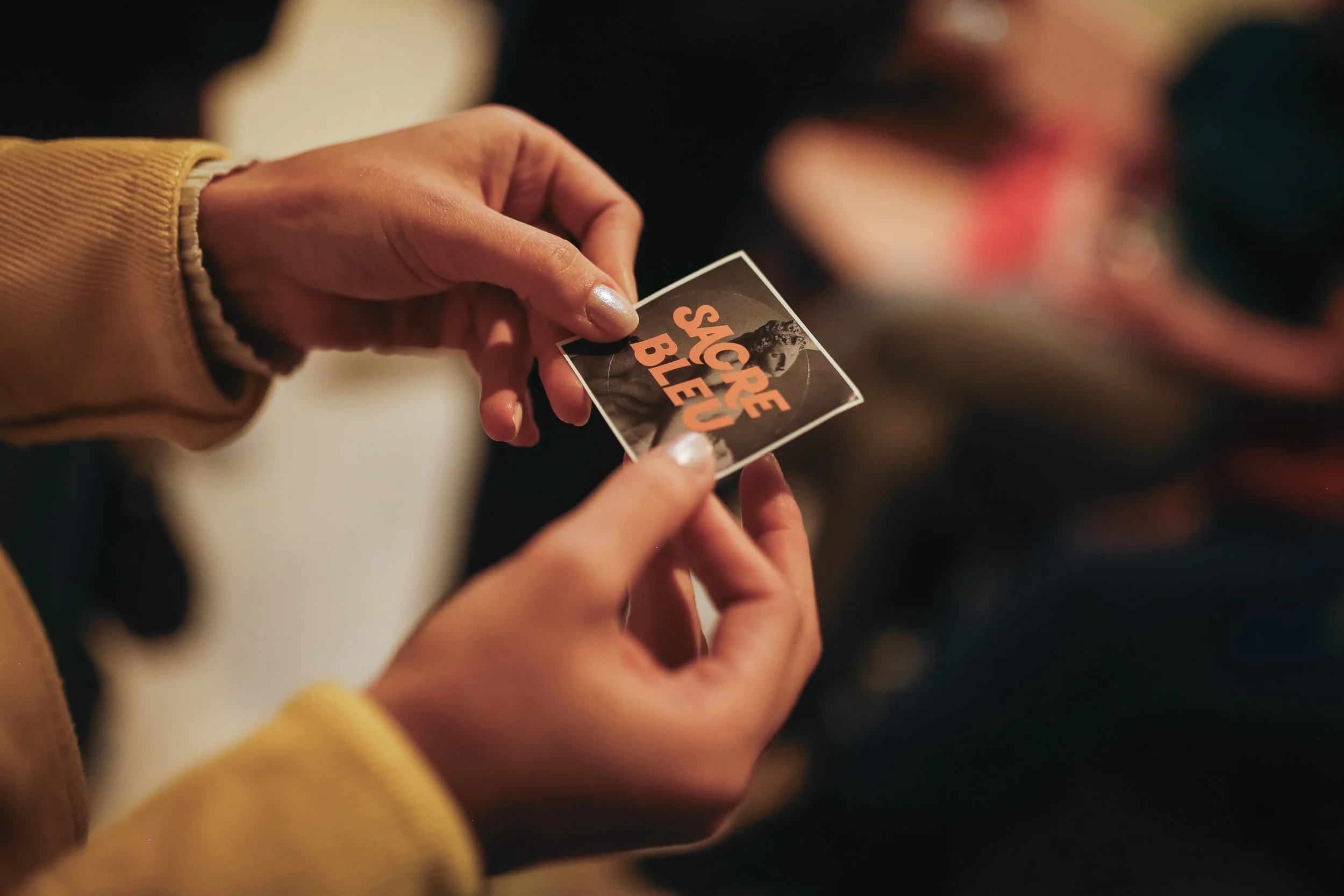 Person holding a square sticker with a black and white image and orange text that reads 'Syrphe Bleu' at an indoor event.