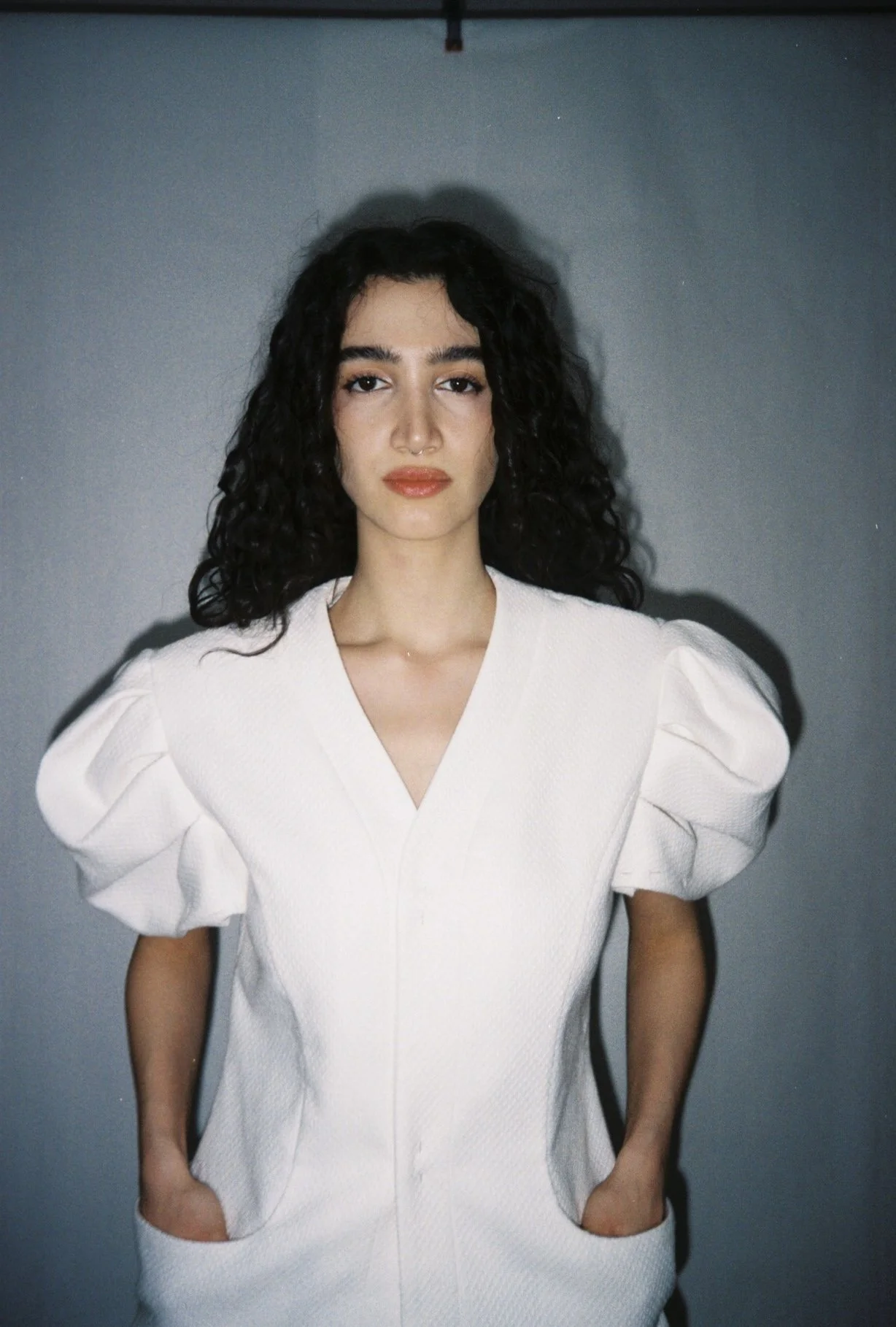 A young woman with curly black hair wearing a white dress with puffed sleeves stands against a gray background.