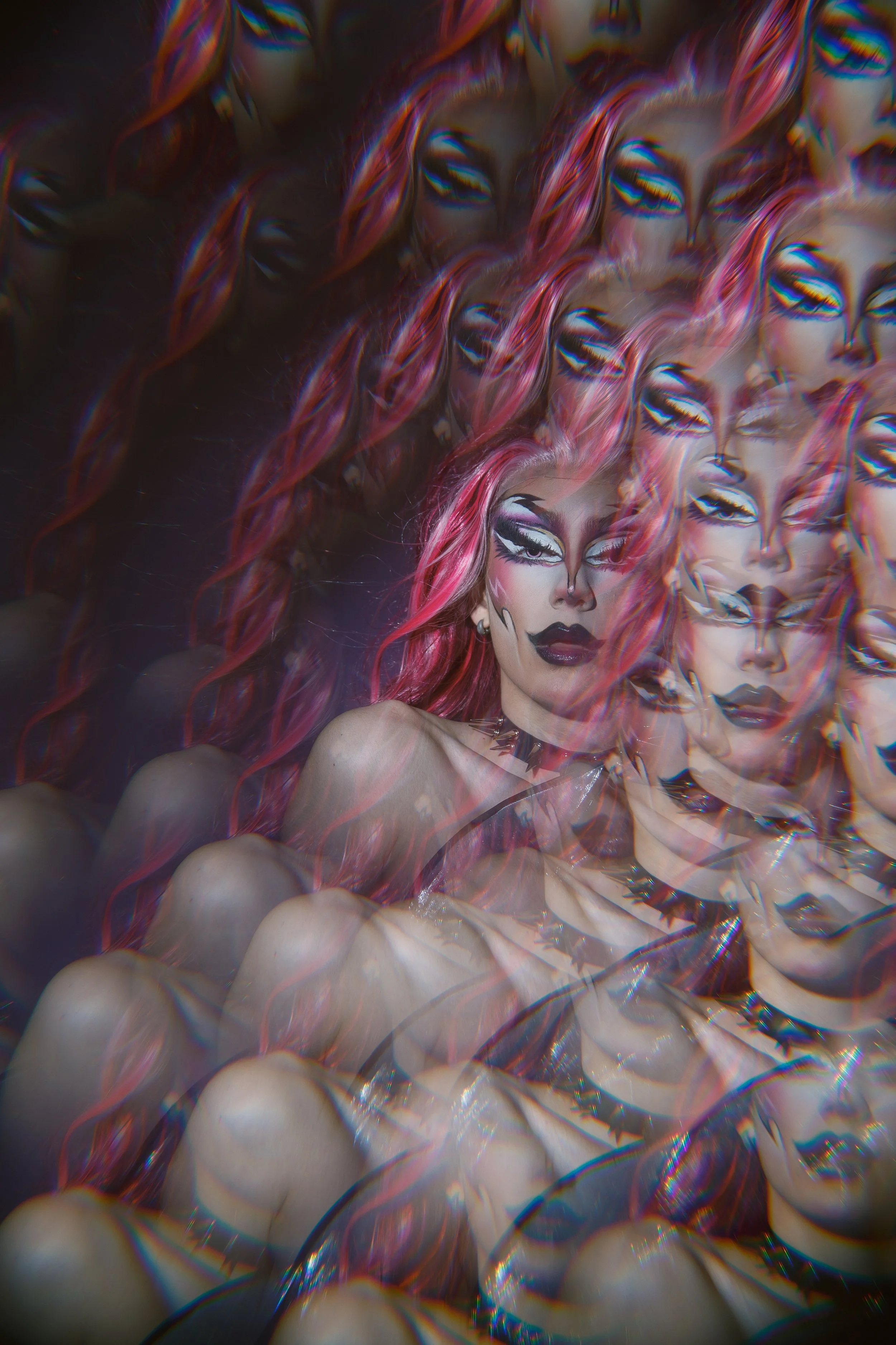 A woman with vibrant pink and orange hair, wearing dark makeup and a spiked choker, is surrounded by multiple overlapping, distorted images of herself creating a kaleidoscope effect.