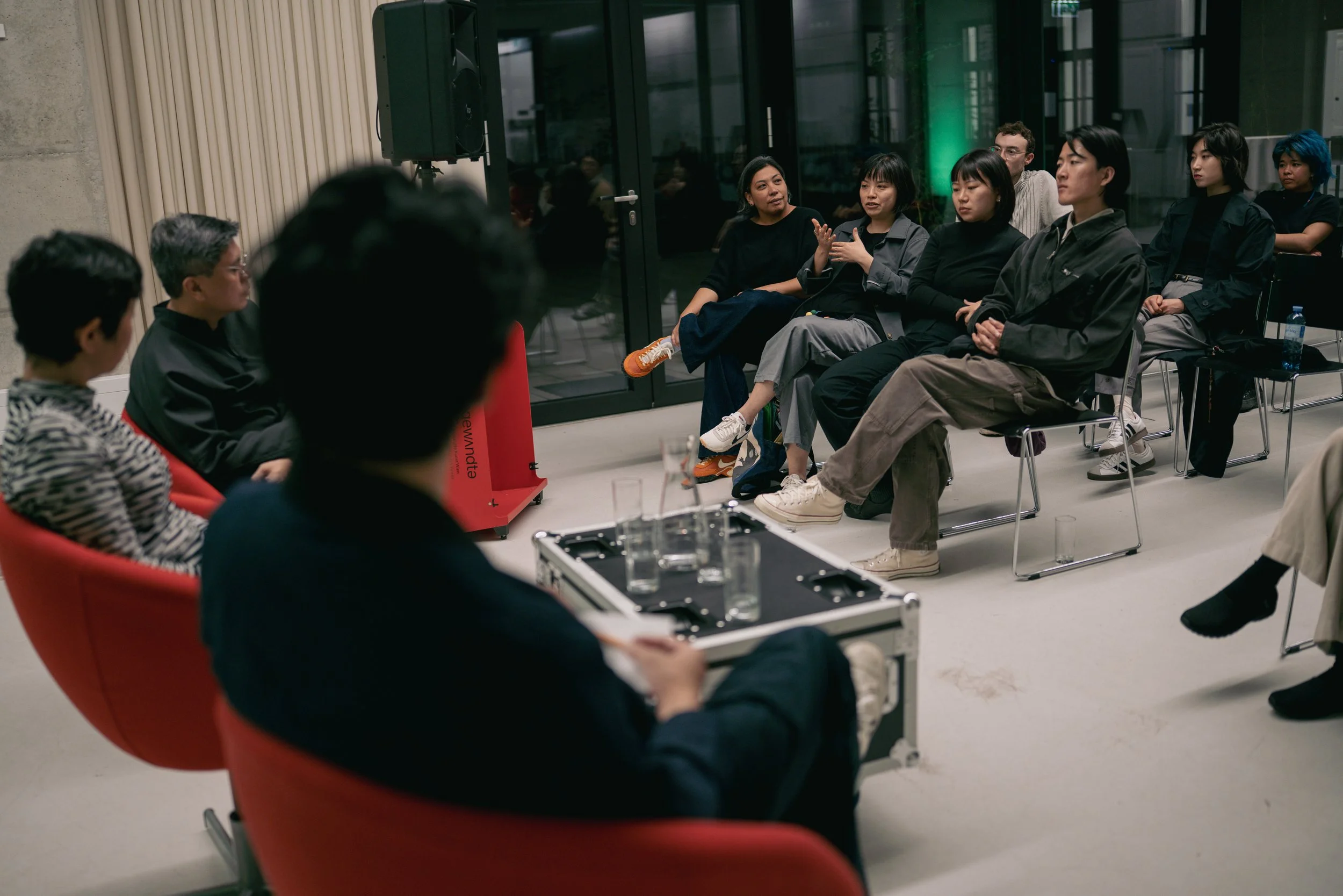 A group of people participating in a panel discussion or meeting in a modern indoor space, with some audience members seated and engaging with the speakers.