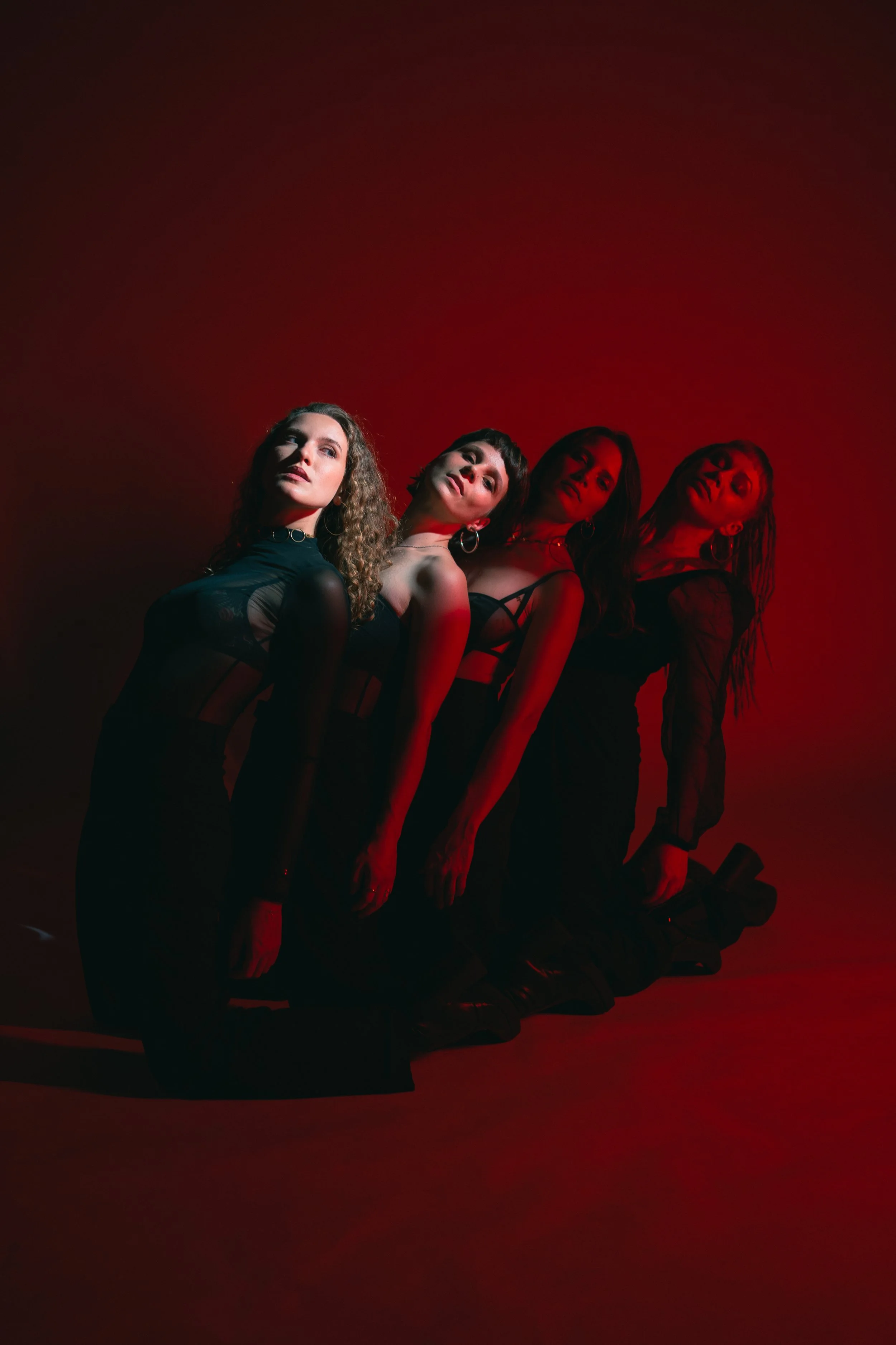 Four women kneeling on the floor in black outfits, illuminated by red lighting, posed against a red background.