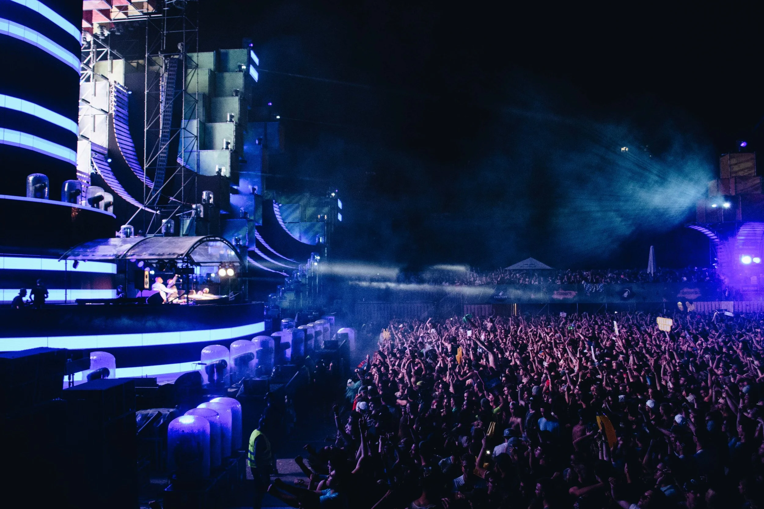 A large crowd at a concert or music festival at night with a brightly lit stage and DJ performing.