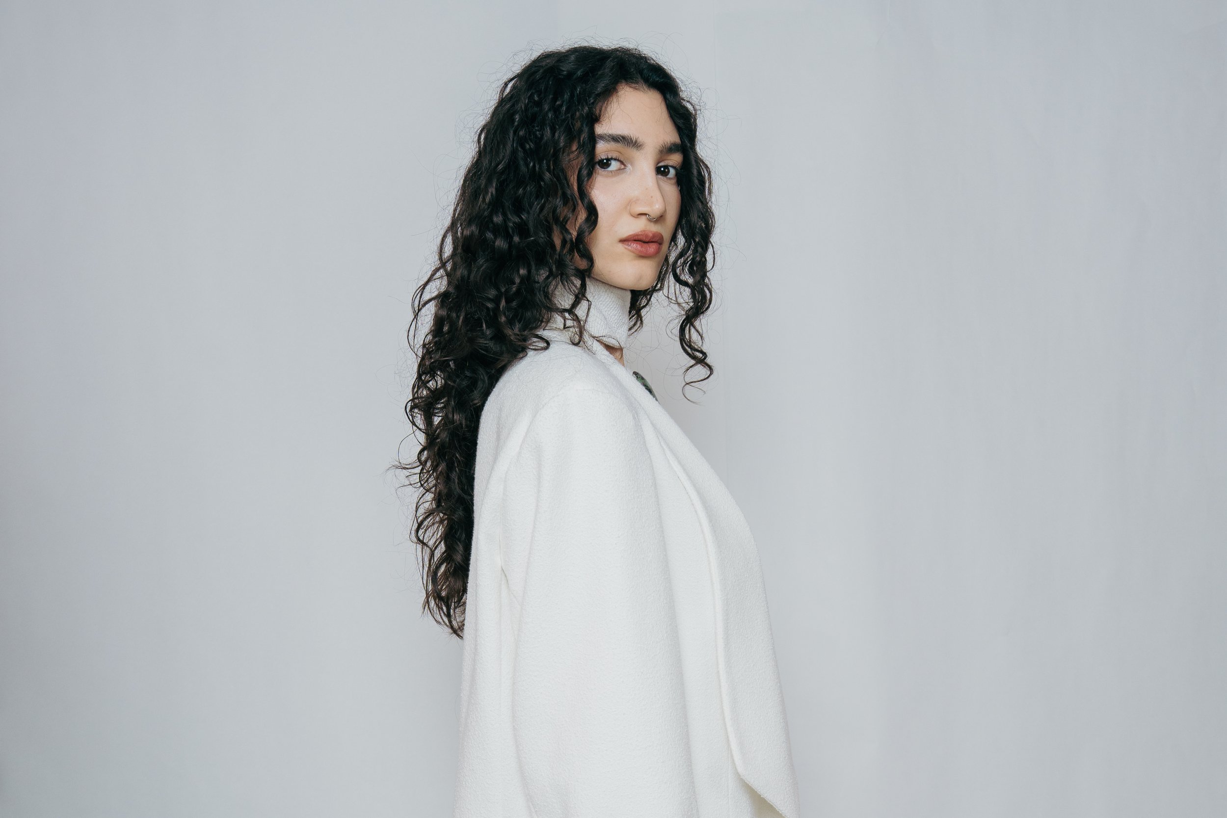 A woman with long, dark curly hair wearing a white coat, looking over her shoulder against a plain gray background.