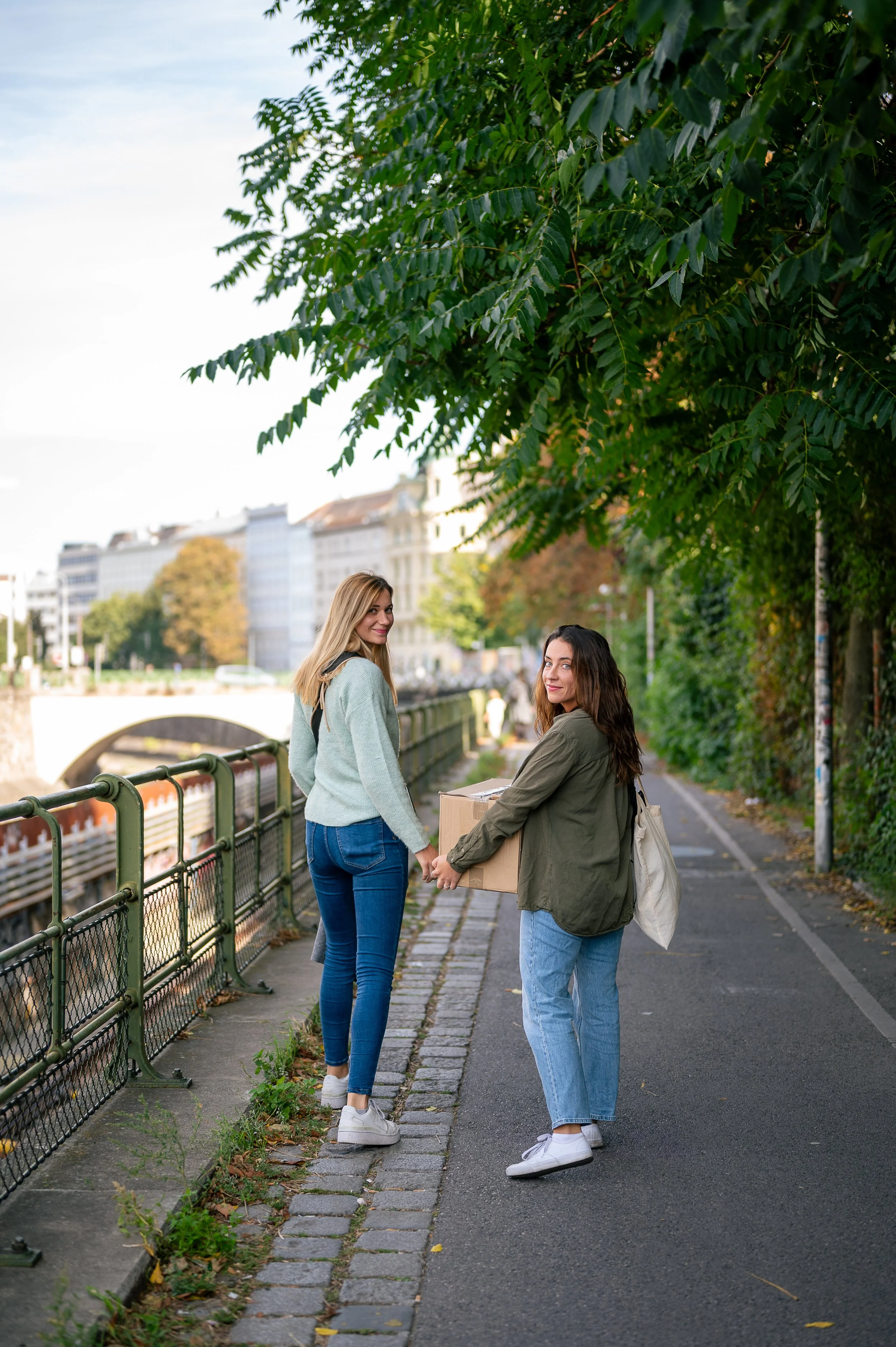 Two women exchanging a cardboard box on the sidewalk near a river, in an urban park setting with trees and buildings in the background.