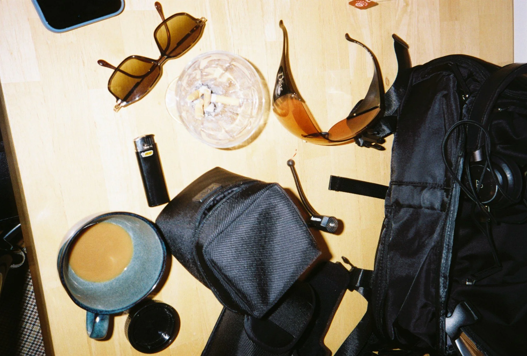 A wooden table with sunglasses, a glass ashtray with cigarette butts, a brown glass bottle, a black camera case, a black backpack, a coffee mug, and a pair of black headphones.