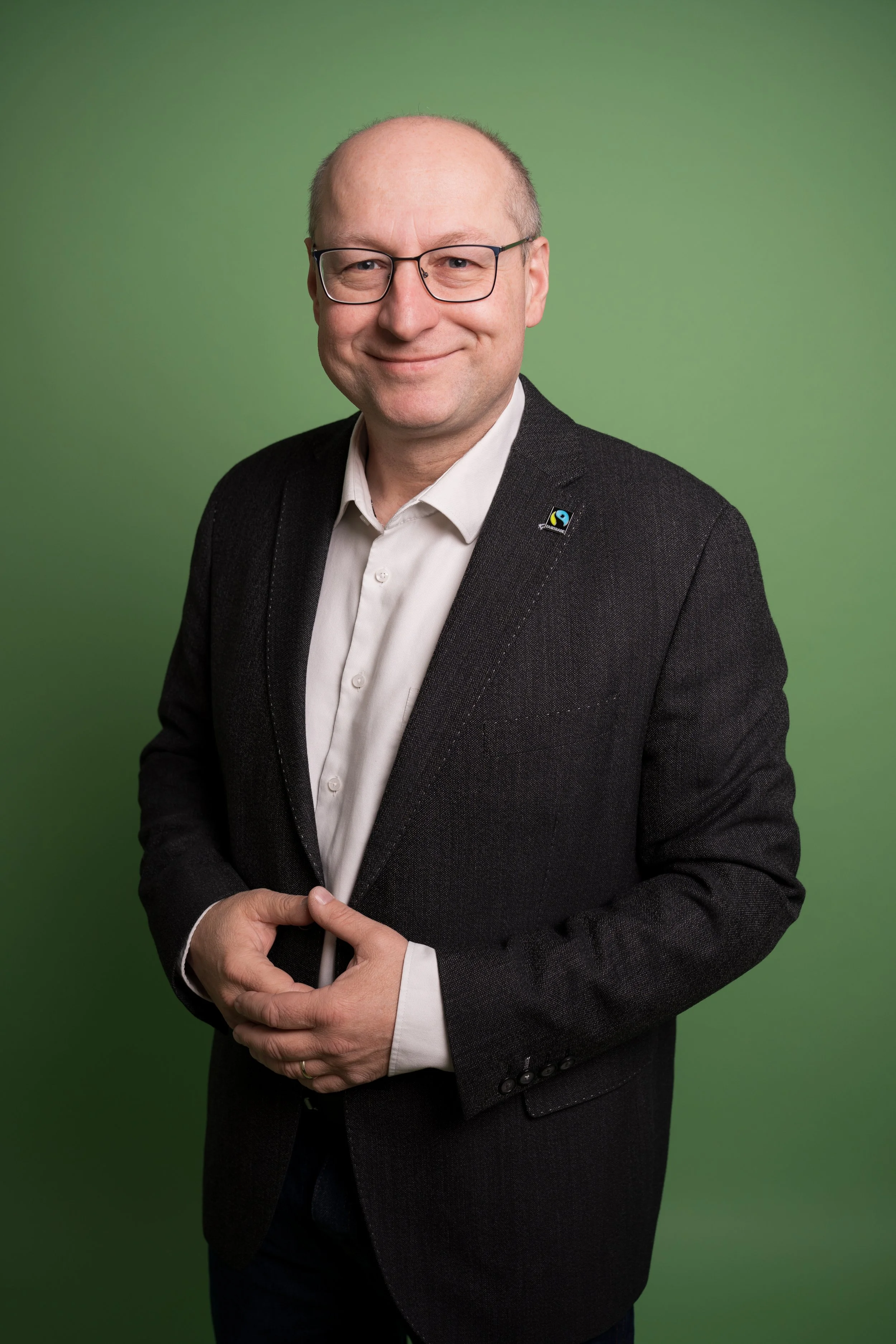Portrait of a smiling man with glasses, wearing a dark suit jacket and a white shirt, standing against a green background.