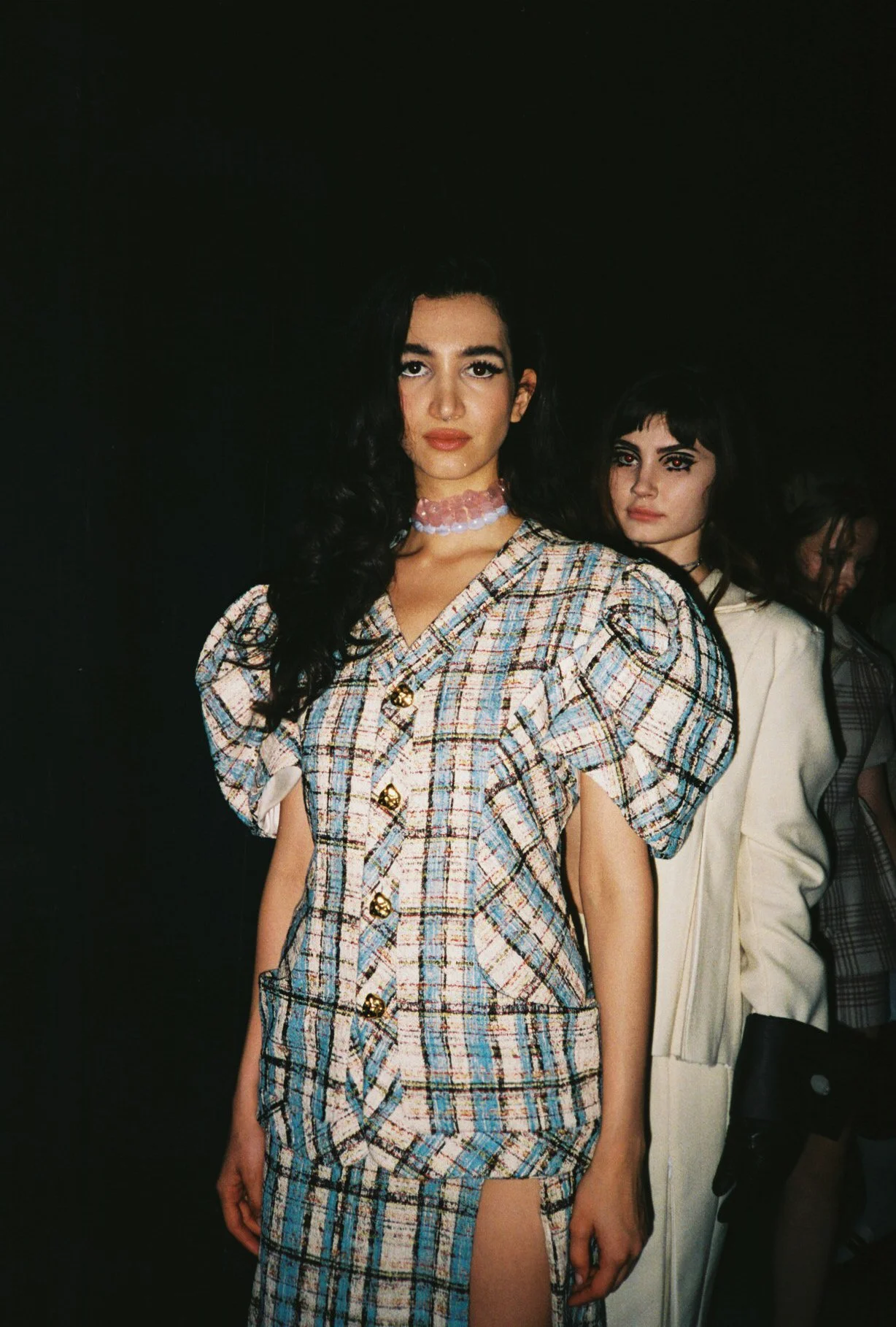Two women standing in front of a dark background, with the woman in the foreground wearing a plaid dress with puffy sleeves, gold buttons, and a slit, and the woman behind her wearing a white coat with a plaid shirt underneath.