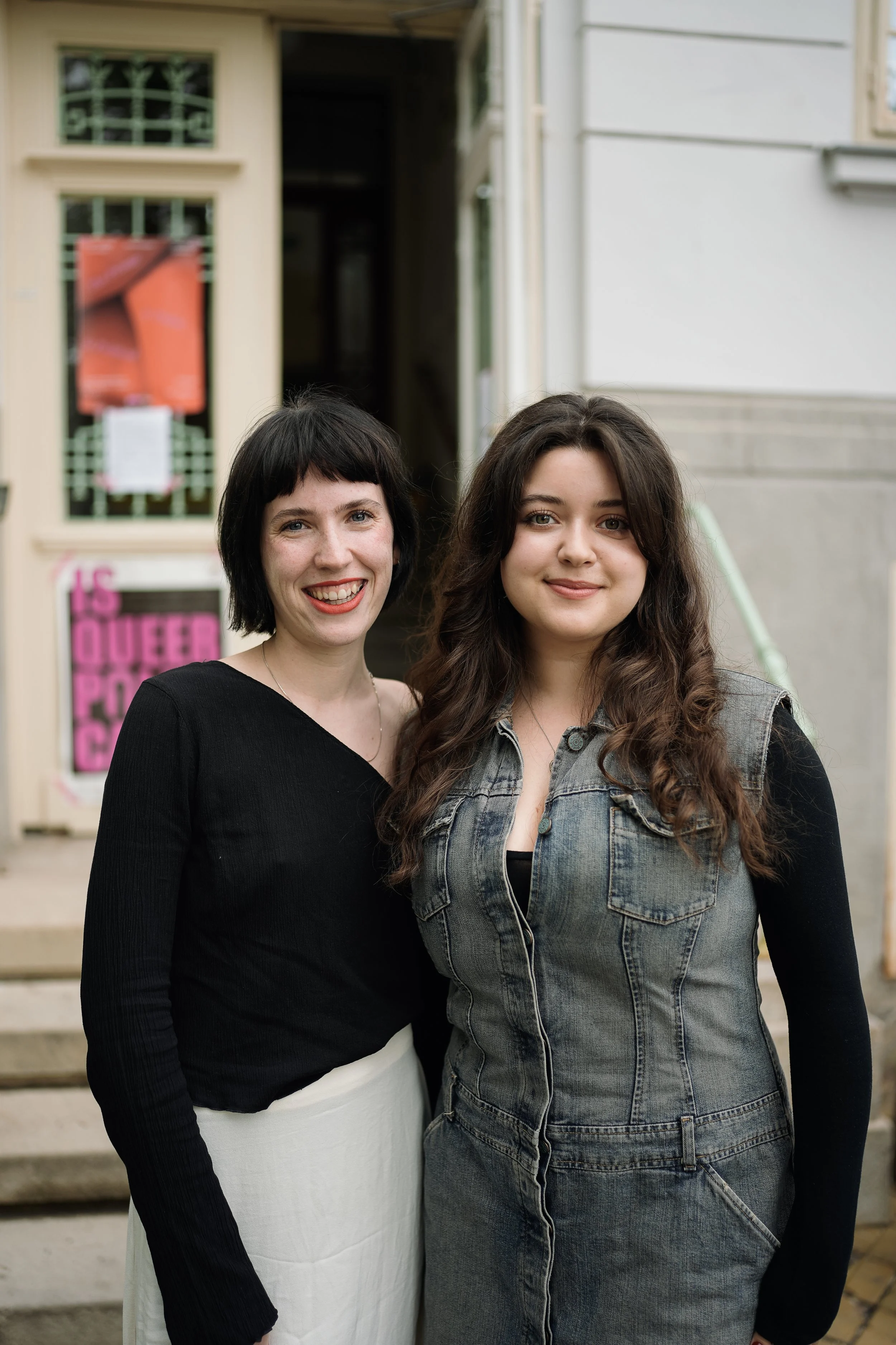 Two women standing outdoors in front of a building, smiling at the camera.
