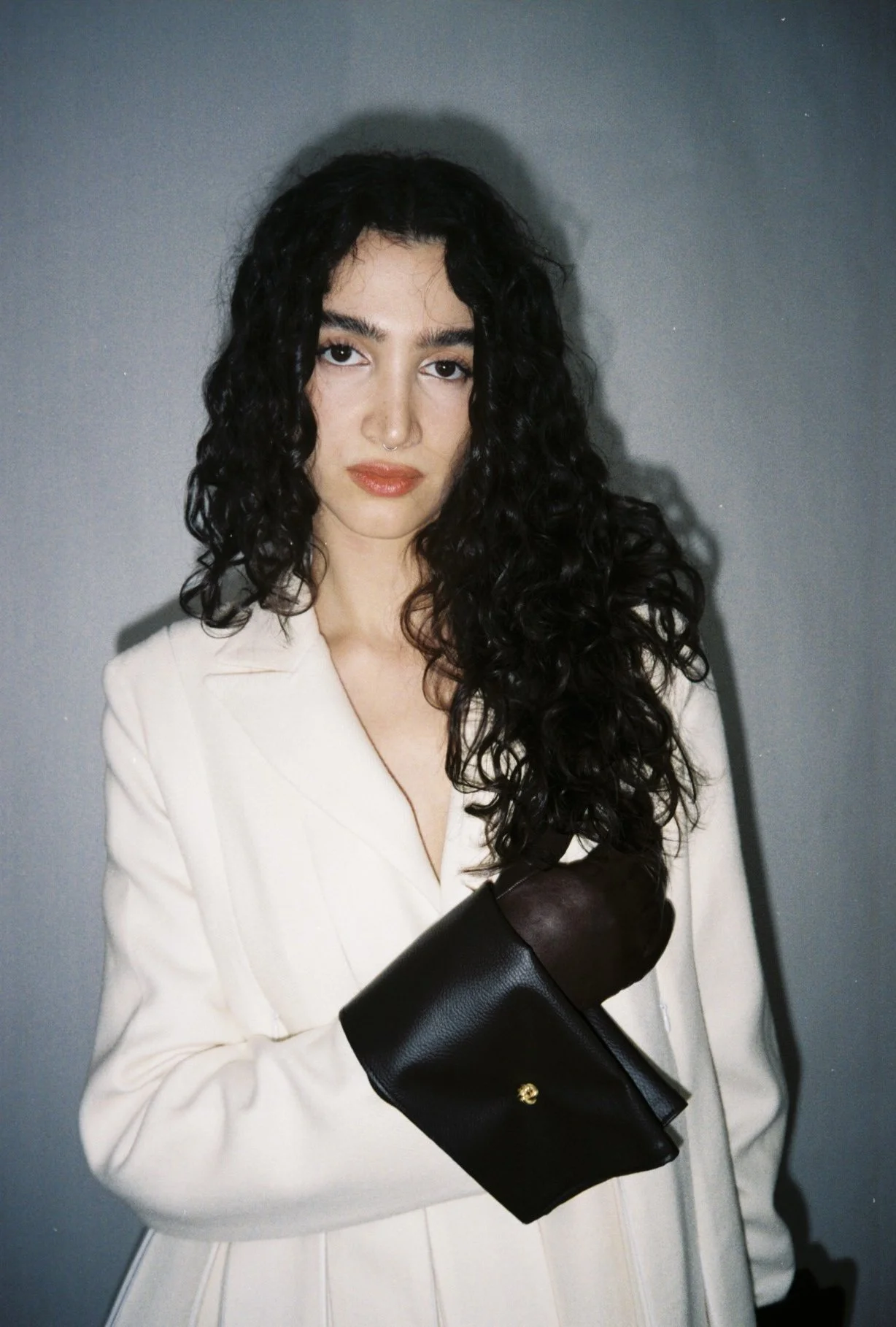 A woman with curly dark hair and fair skin stands against a gray background. She is wearing a cream-colored blazer with pleats and a black leather glove on her right hand, which she holds close to her chest. She has a nose ring and wears makeup with 