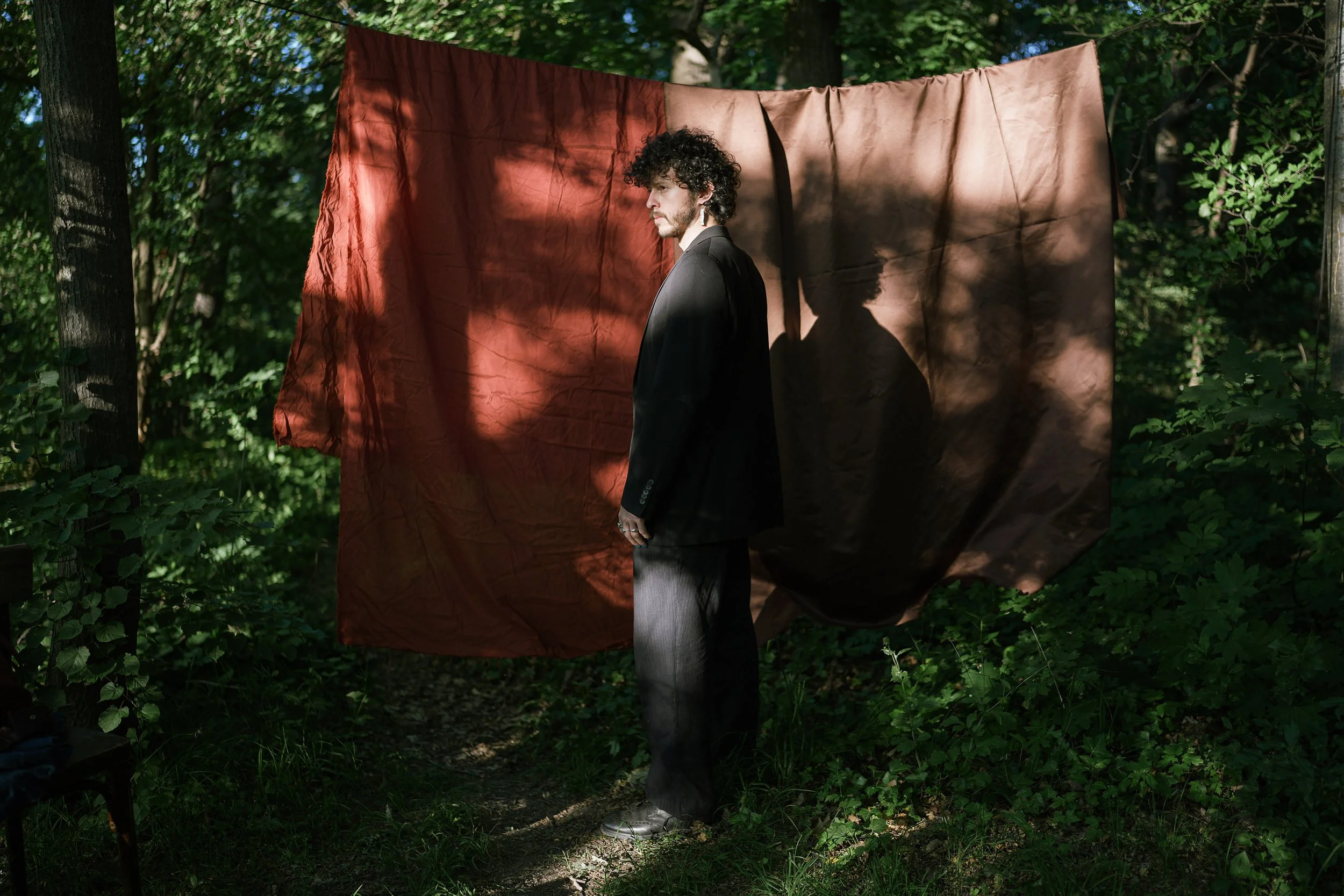 A man in a black suit stands outdoors in front of a brown cloth backdrop, with sunlight casting shadows on the backdrop and his face.