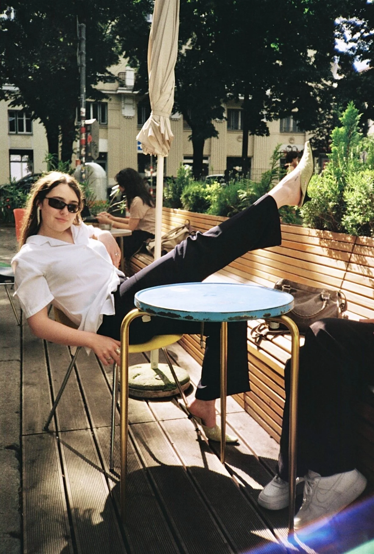 A woman with sunglasses wearing a white blouse and black pants, sitting outdoors at a cafe table, raising her leg on a bench, with trees and other people in the background.
