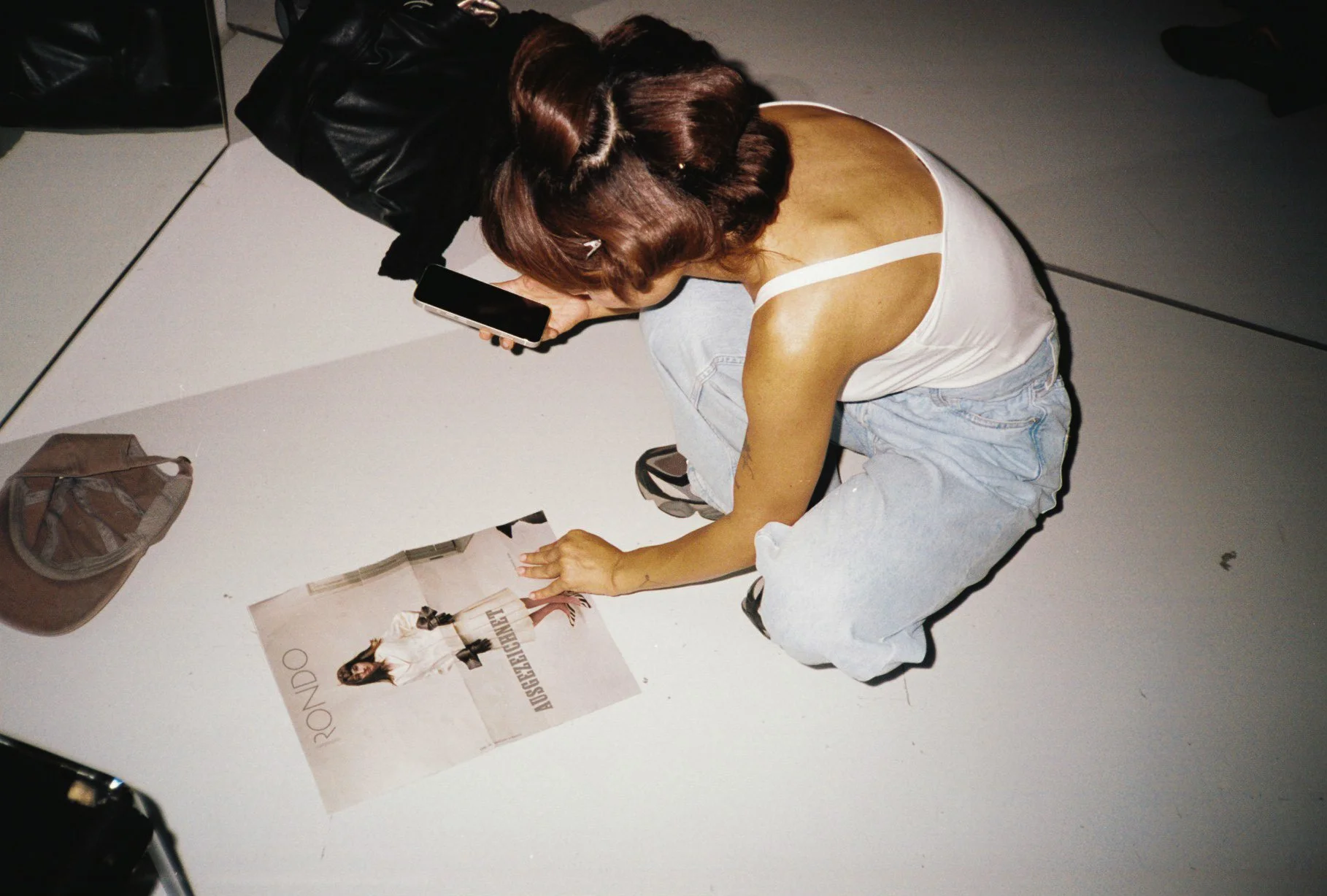 A woman with brown hair styled in vintage waves is sitting on the floor, looking at her phone, beside a poster on the ground. She is wearing a white tank top and light blue jeans.
