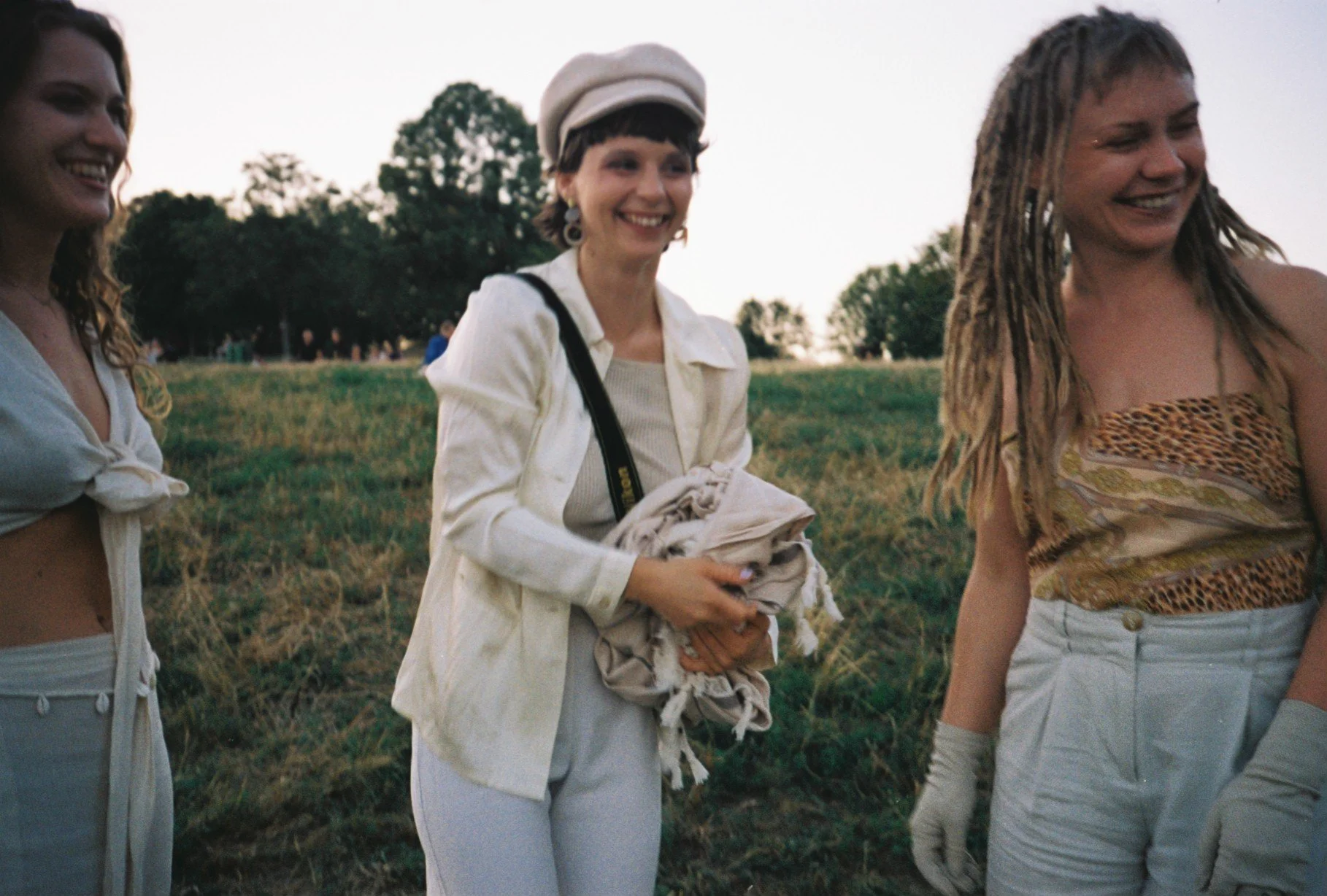 Three women standing outdoors in a grassy field during sunset, smiling and enjoying themselves.