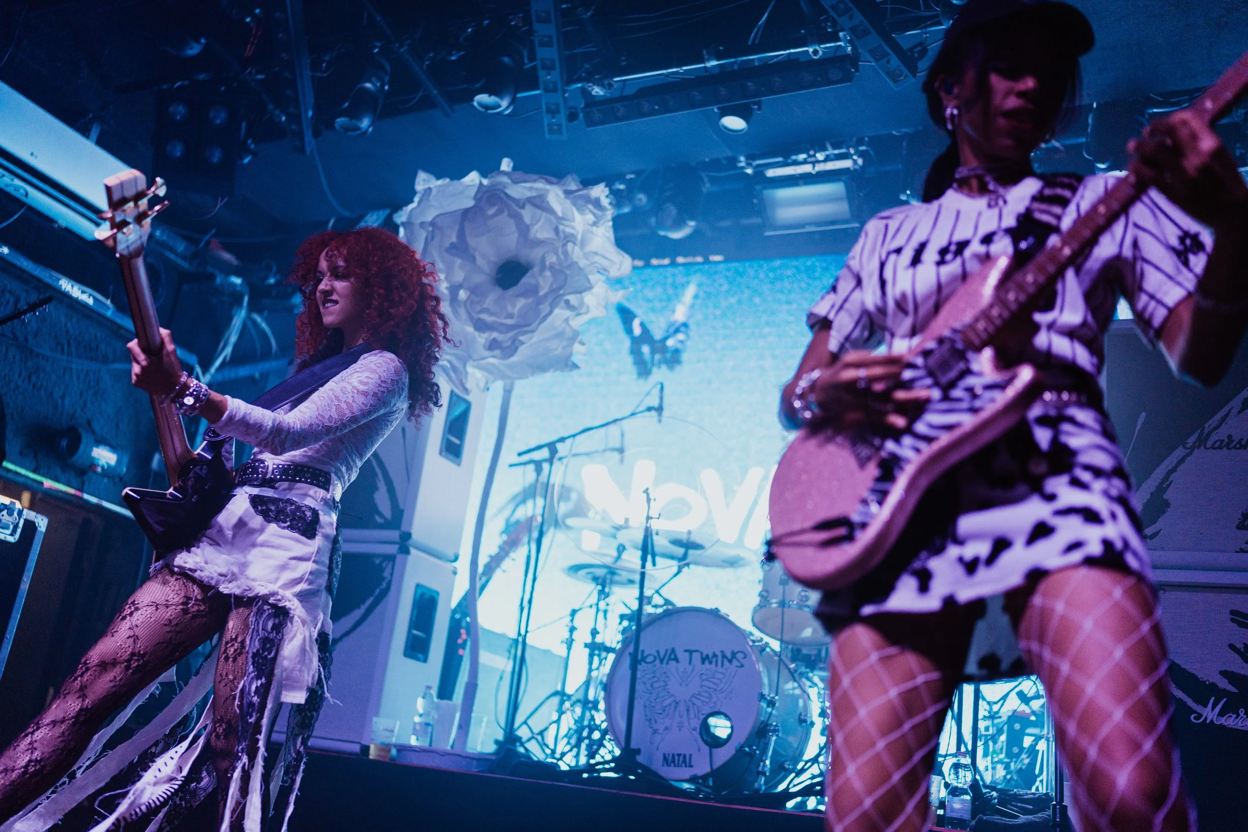 Two women perform on stage with electric guitars, wearing fishnet tights and fashionable outfits, with musical instruments and a drum set in the background.