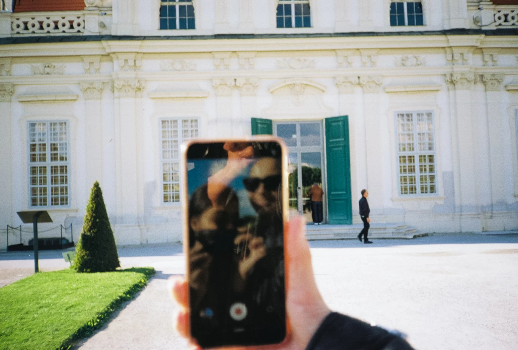 Person taking a photo of a building using a smartphone, with the phone screen showing a face with sunglasses, while the building has large green doors and windows.