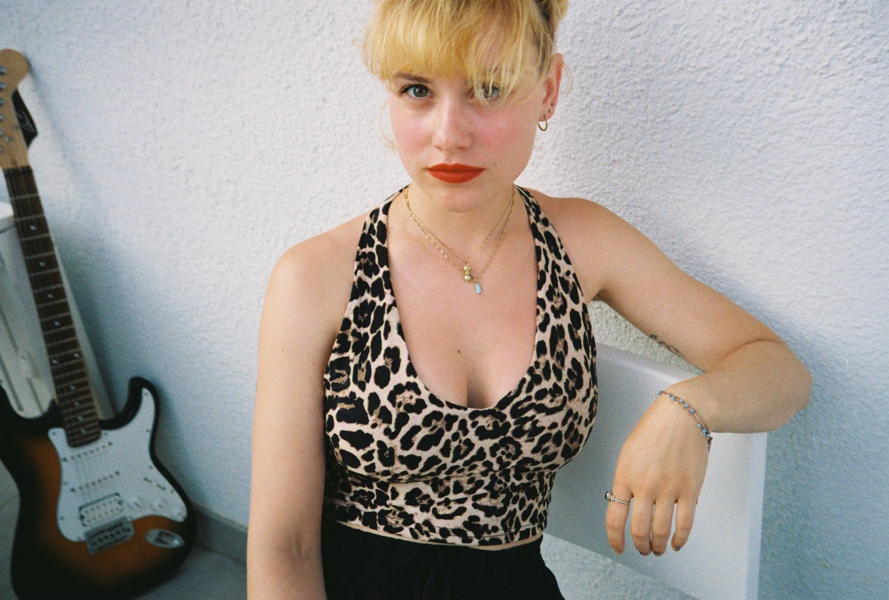 A young woman with blonde hair and red lipstick wearing a leopard print tank top, jewelry, and earrings sitting against a white wall with a guitar in the background.