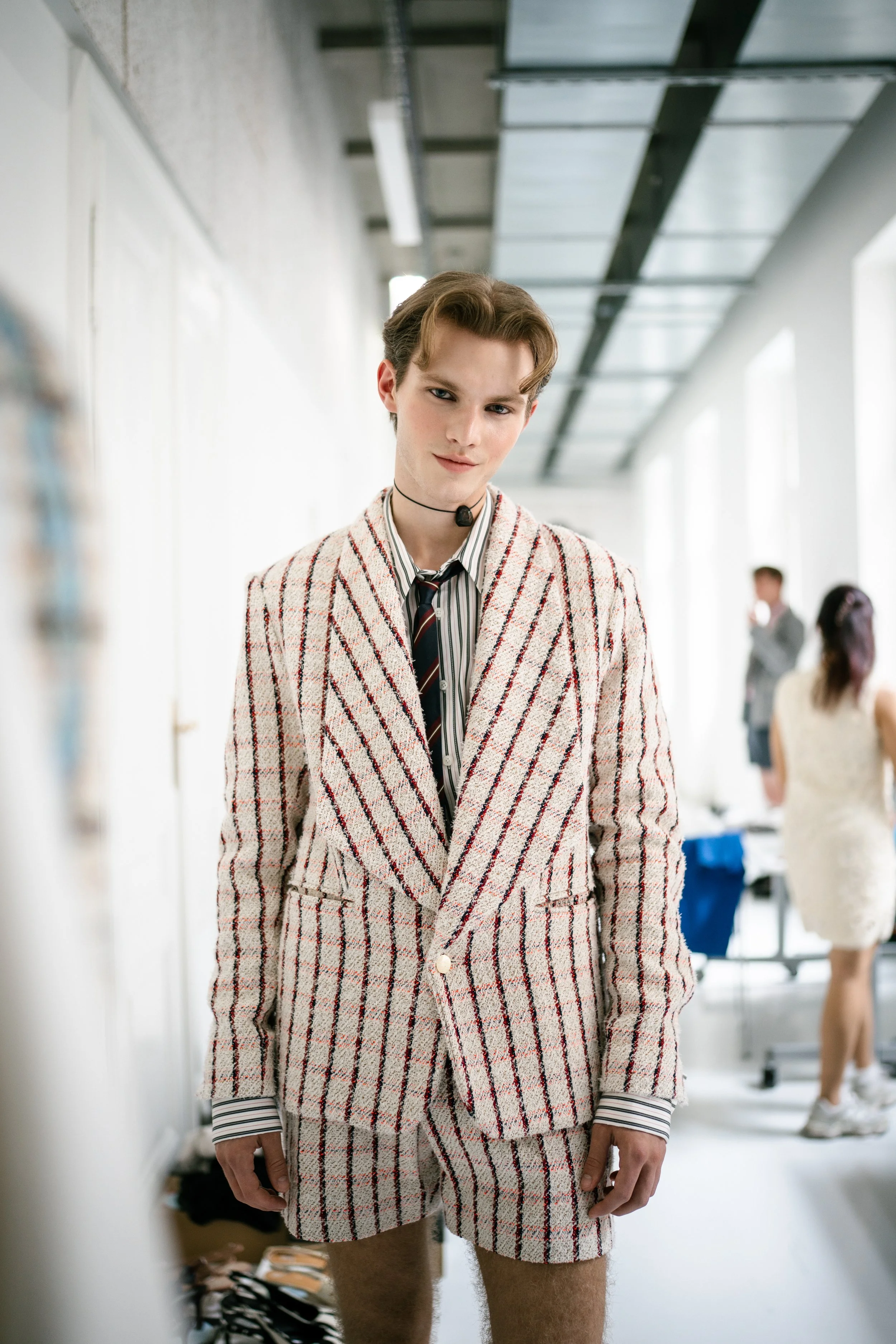 Young man with styled hair wearing a striped dress shirt, a striped tie, a plaid blazer, and matching plaid shorts, standing in a well-lit room with other people in the background.