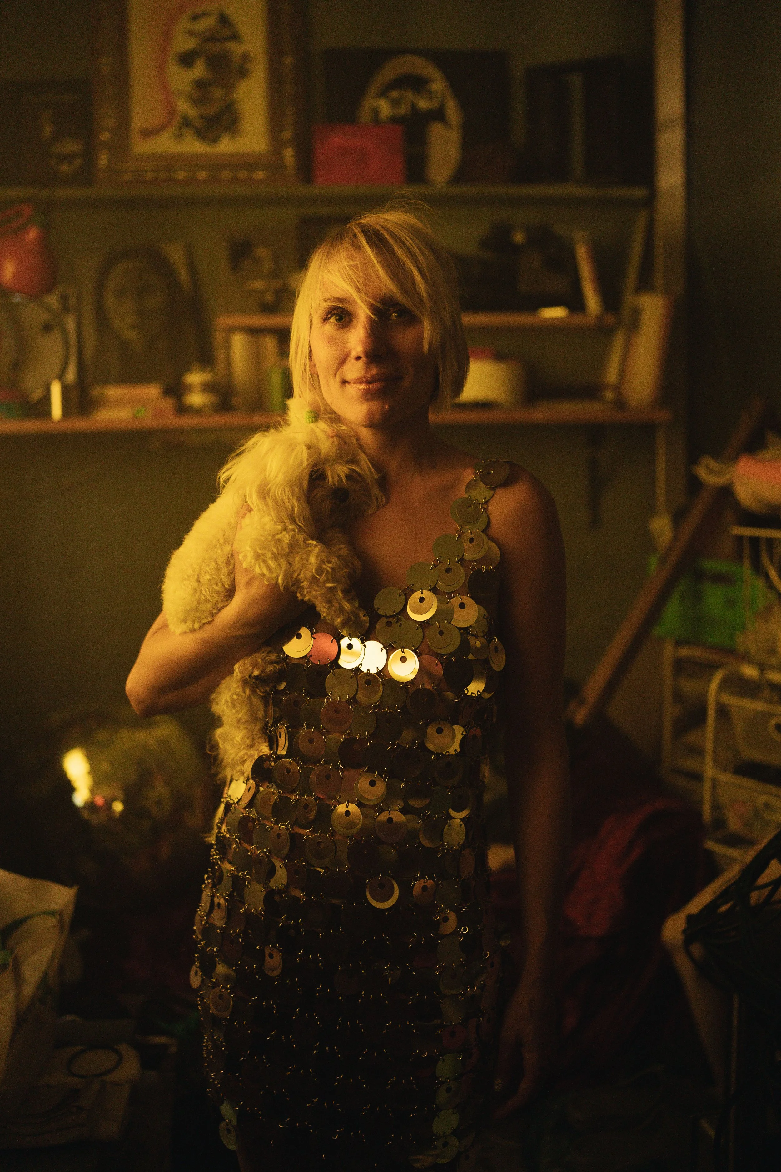 A woman with blonde hair holding a small tan Poodle dog, standing in a cluttered room with books, art, and shelves in the background, dressed in a shiny, sequined dress.