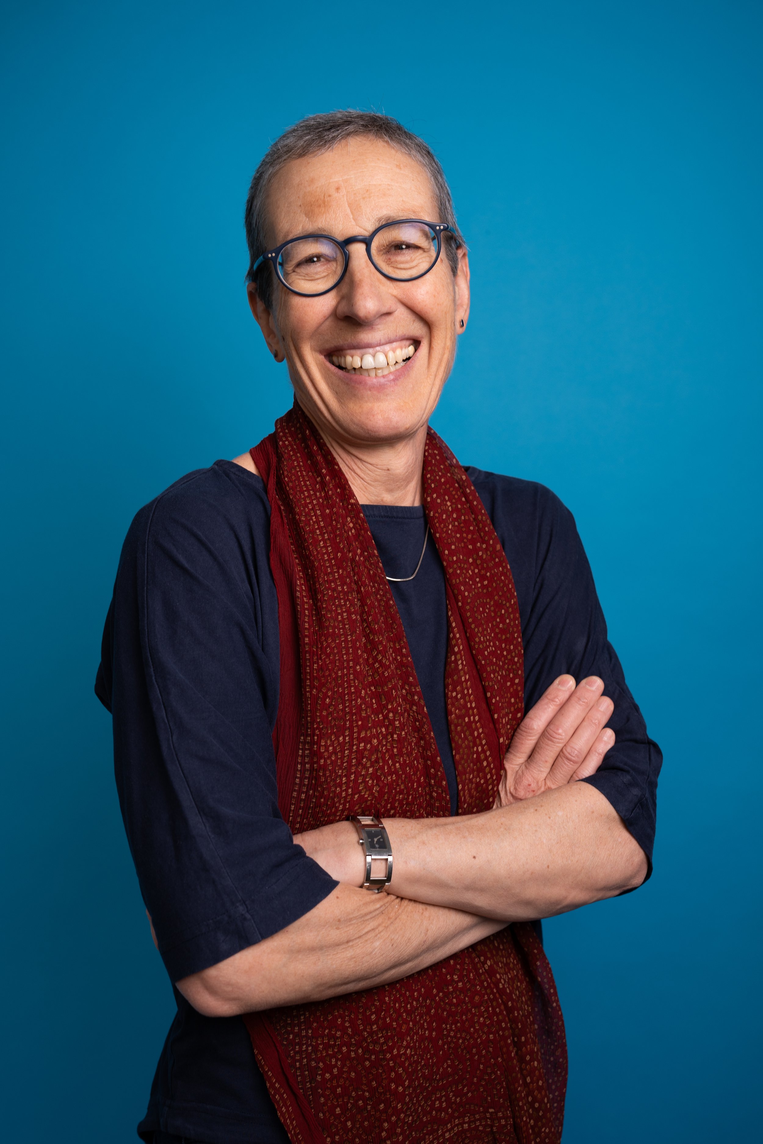 A smiling woman with short gray hair, glasses, and earrings, wearing a navy shirt and a red patterned scarf, standing with arms crossed against a blue background.