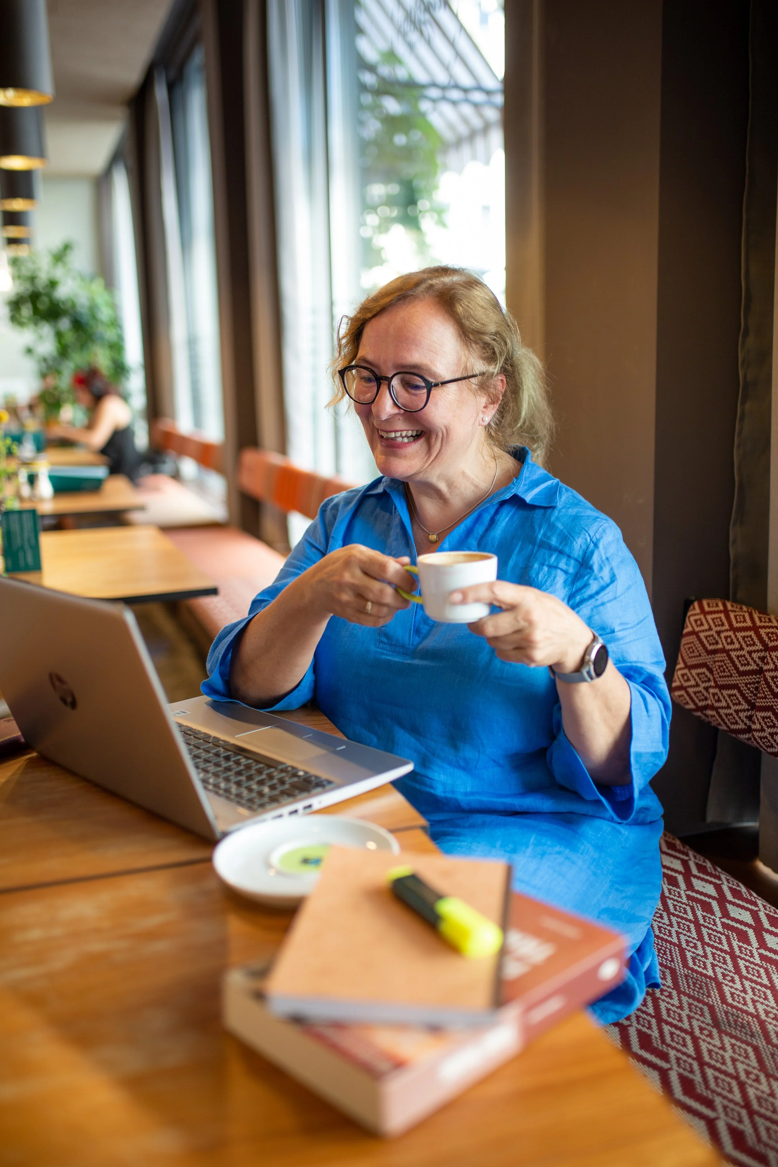 A woman with glasses and a blue shirt smiling while holding a coffee cup, sitting at a wooden table with a laptop, notebooks, a highlighter, and a plate in a cozy cafe with large windows.