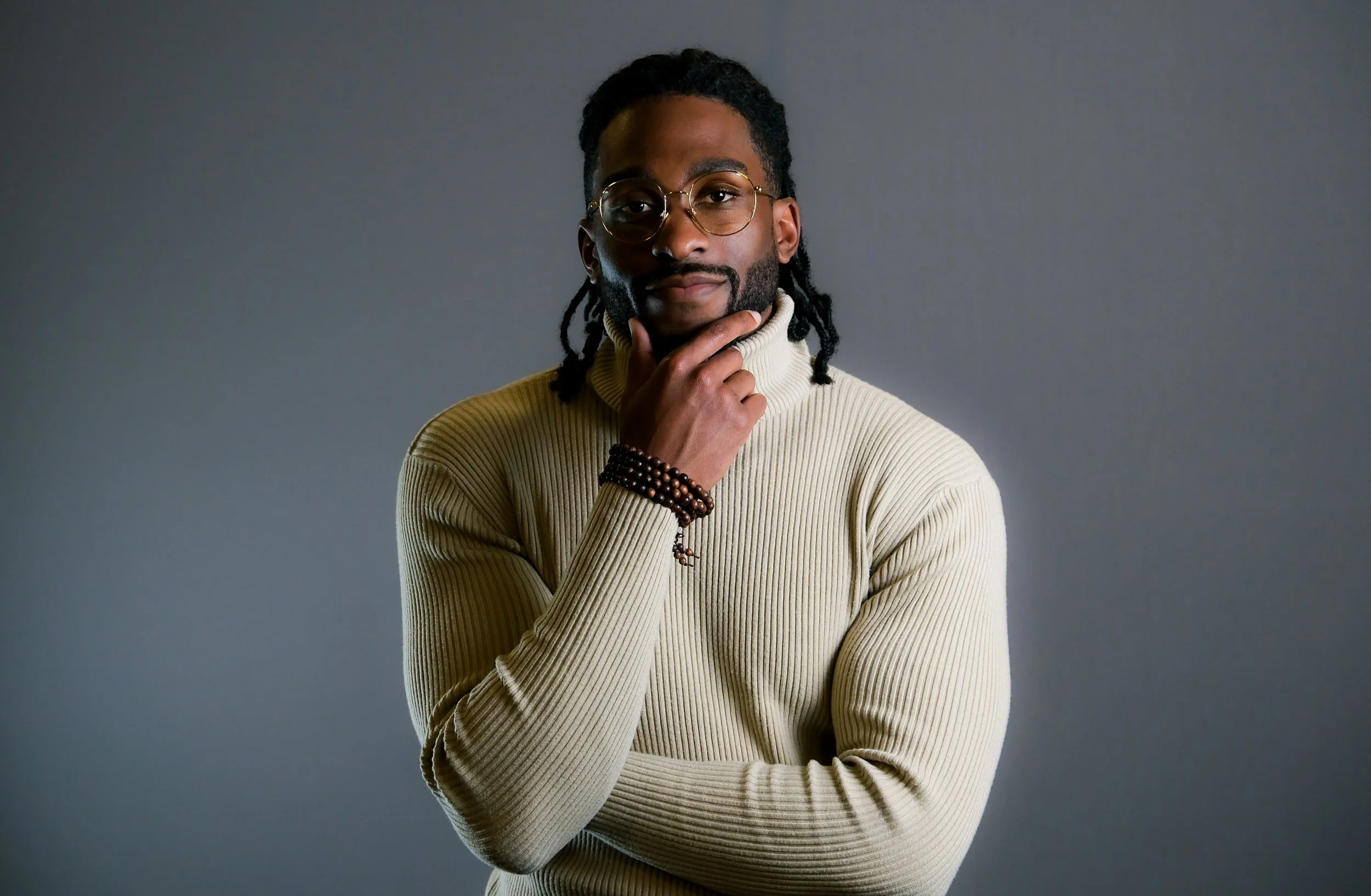 A man with glasses and dreadlocks, wearing a cream-colored turtleneck sweater, stands with one hand touching his chin against a plain gray background.