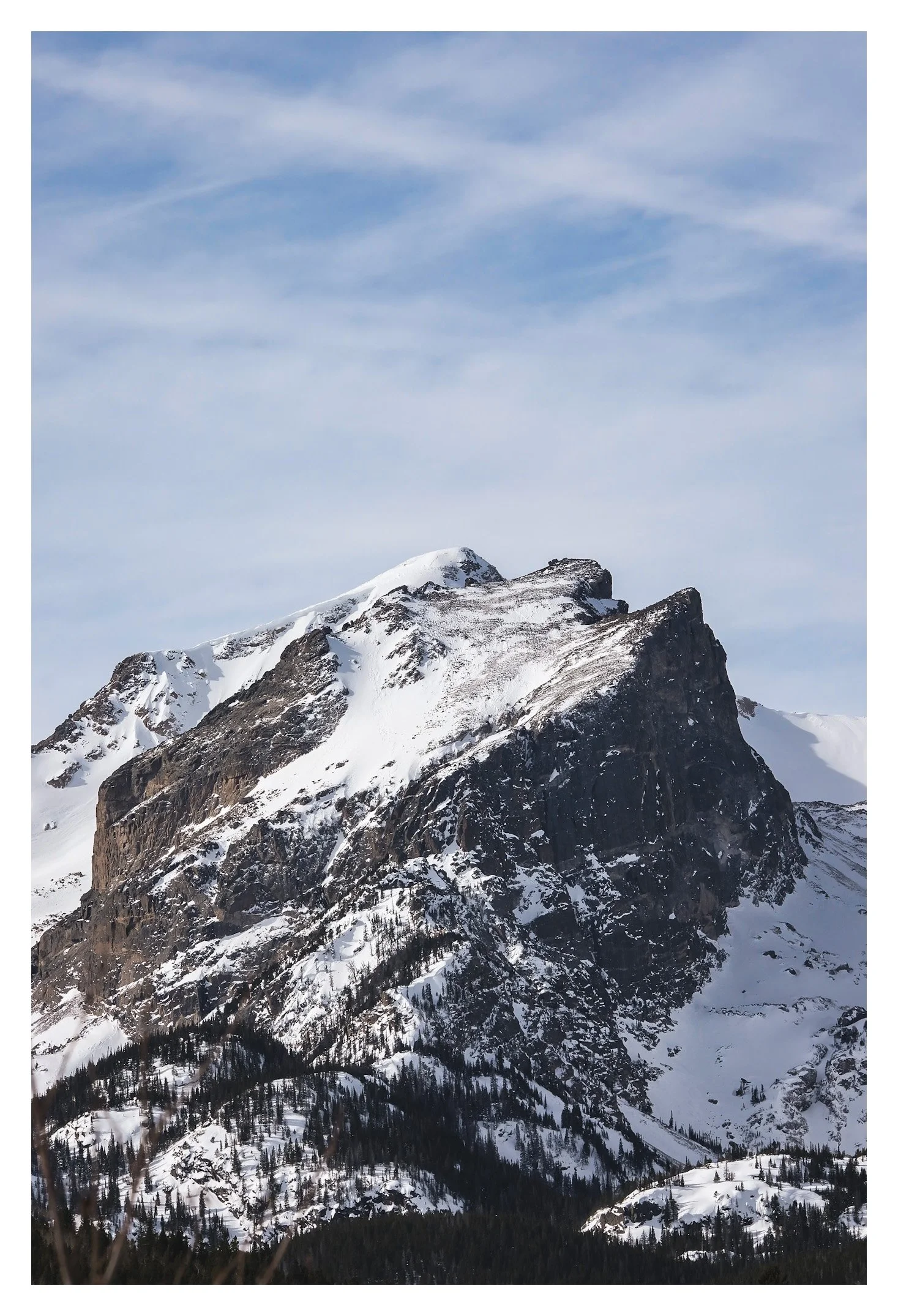 Snow-covered mountain peak with dark rocky faces, below a forested slope with snow, under a blue sky with wispy clouds.