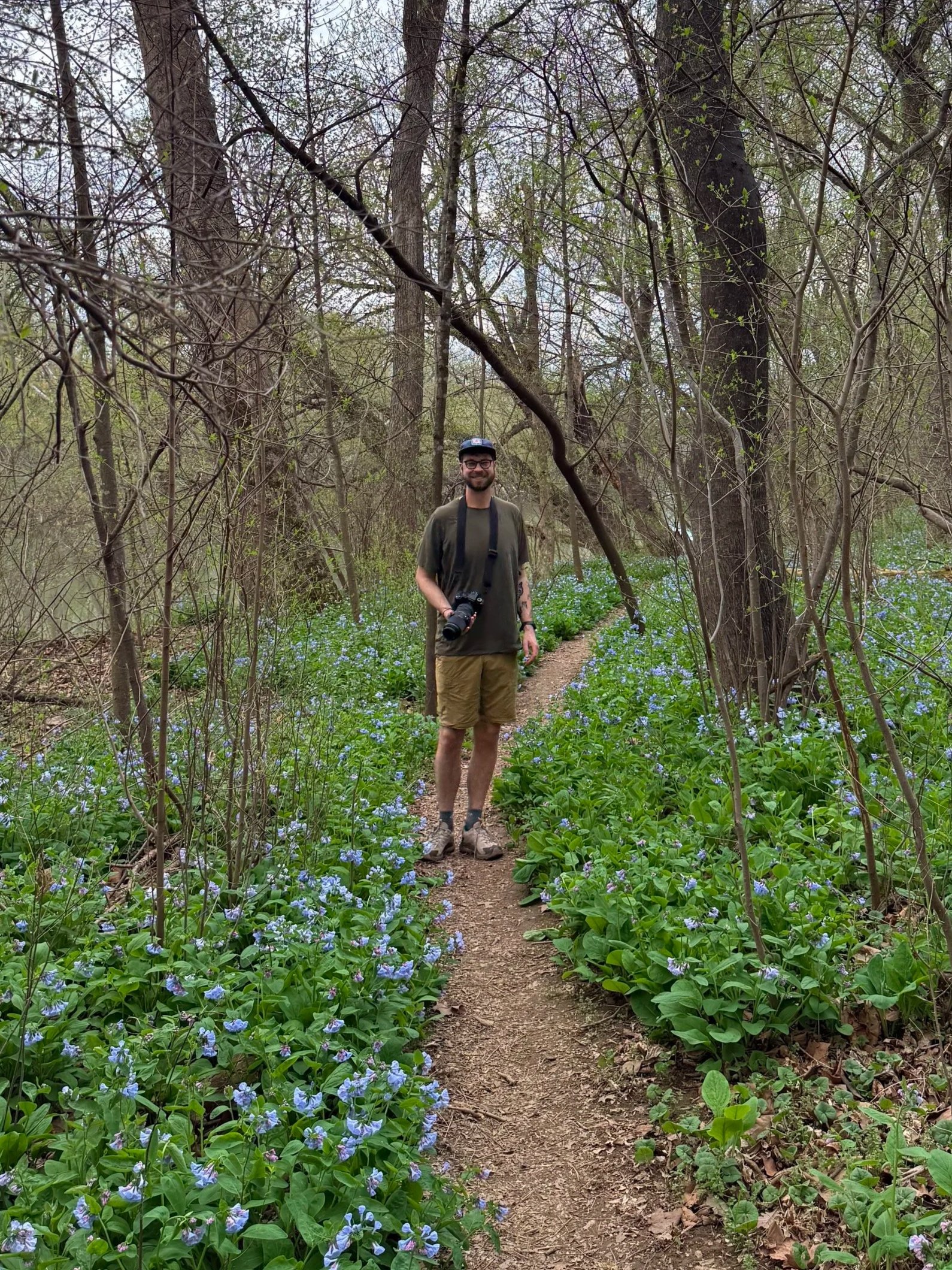 A man smiling and holding a camera stands on a dirt path in a forest surrounded by green foliage and purple flowers.