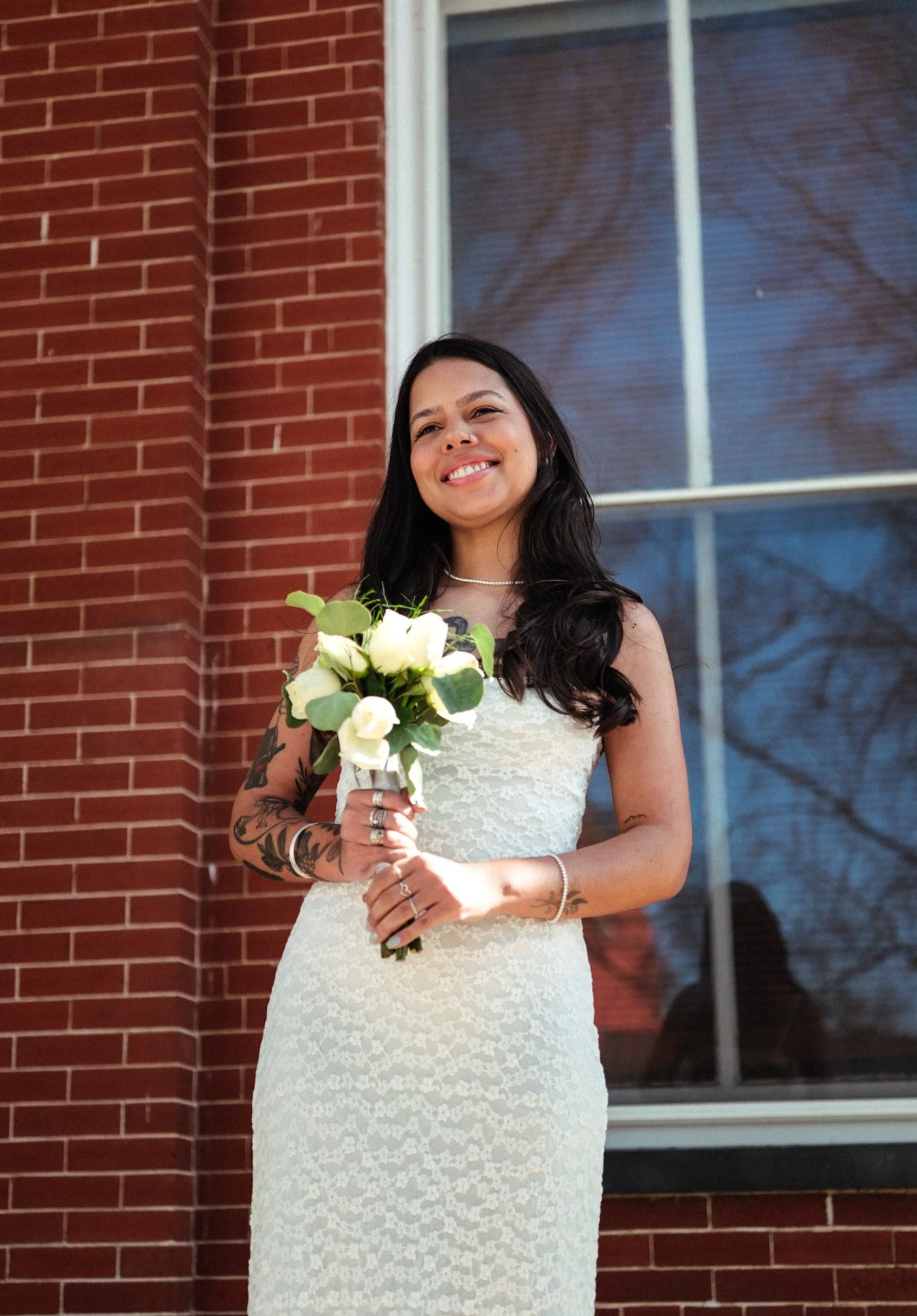 Woman with long dark hair wearing a white lace wedding dress, holding a bouquet of white roses and greenery, standing in front of a red brick wall and a window with a dark pattern.