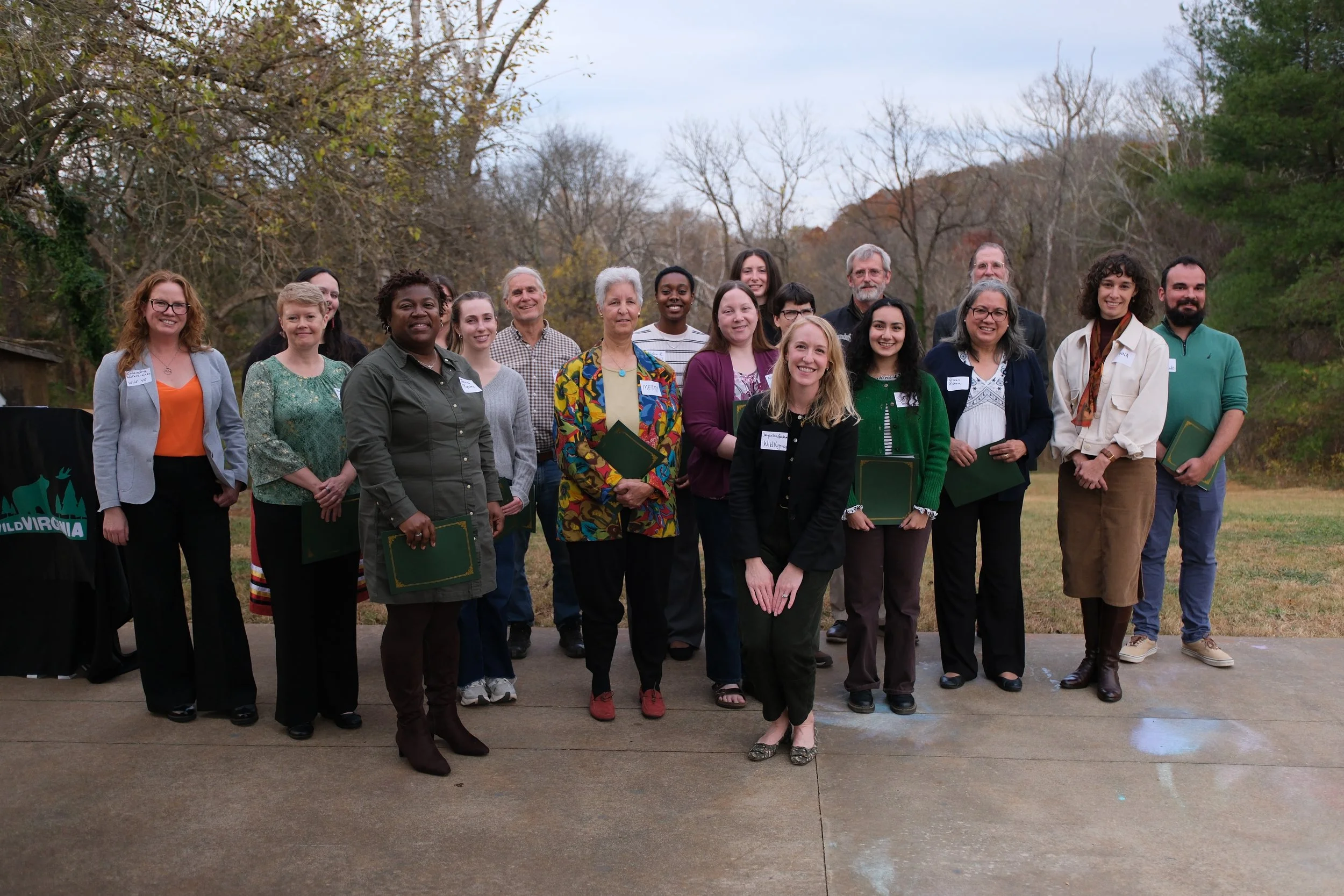 Group of diverse people standing outdoors in a park, posing for a photo. They are dressed in casual and semi-formal clothing, and some are holding documents. Trees with autumn leaves are visible in the background.