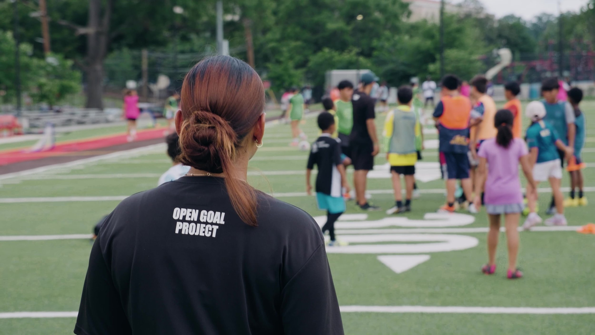 A woman with brown hair in a ponytail, wearing a black shirt with 'Open Goal Project' written on the back, standing on a sports field, observing children and adults participating in a sporting event in the background.