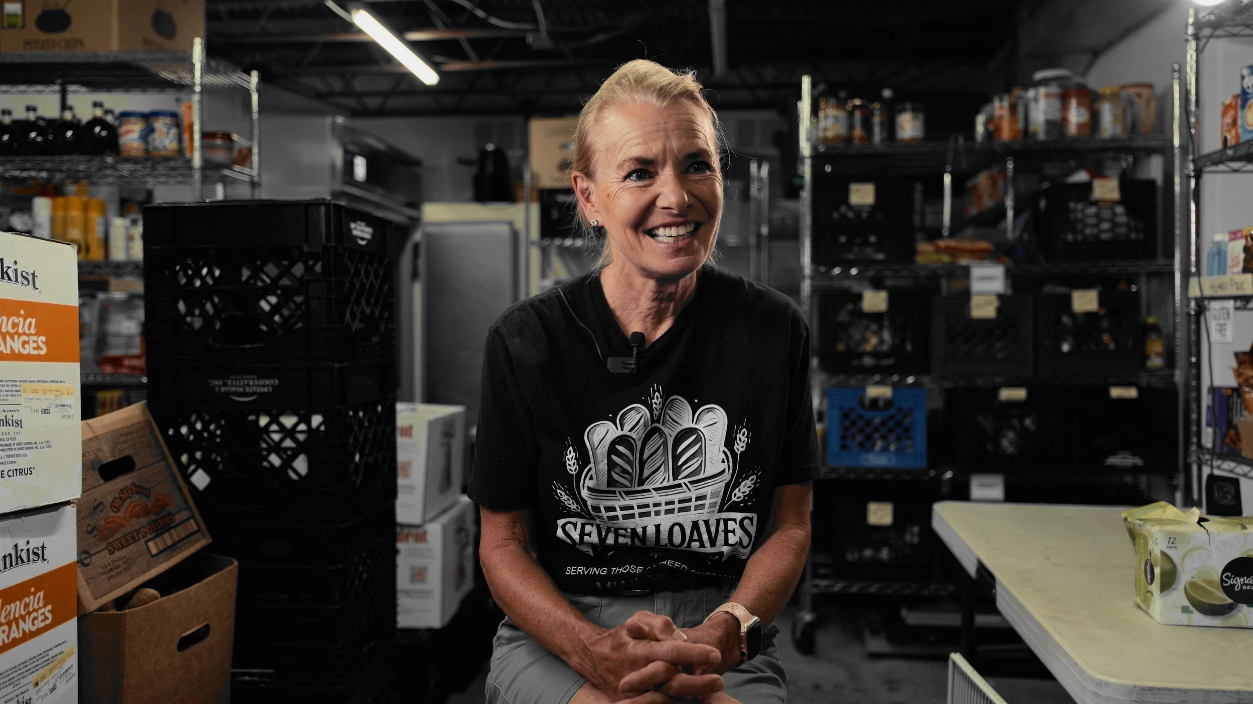 A woman smiling while sitting in a wholesale store surrounded by shelves and boxes of produce and goods. She is wearing a black T-shirt with a bread basket graphic and the words "Seven Loaves."