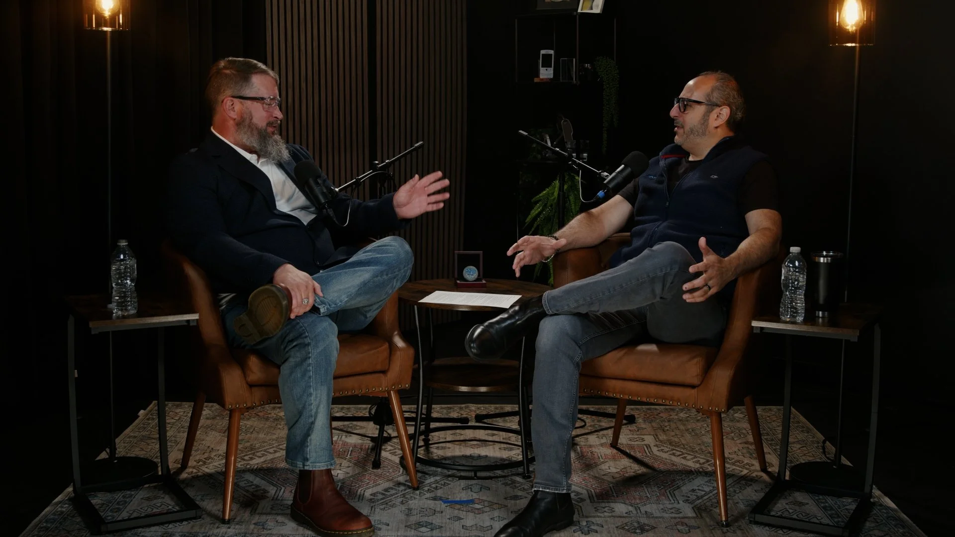 Two men having a conversation in a podcast studio, sitting across from each other with microphones, water bottles, and a small table between them, in a dimly lit room with dark walls and warm lighting.