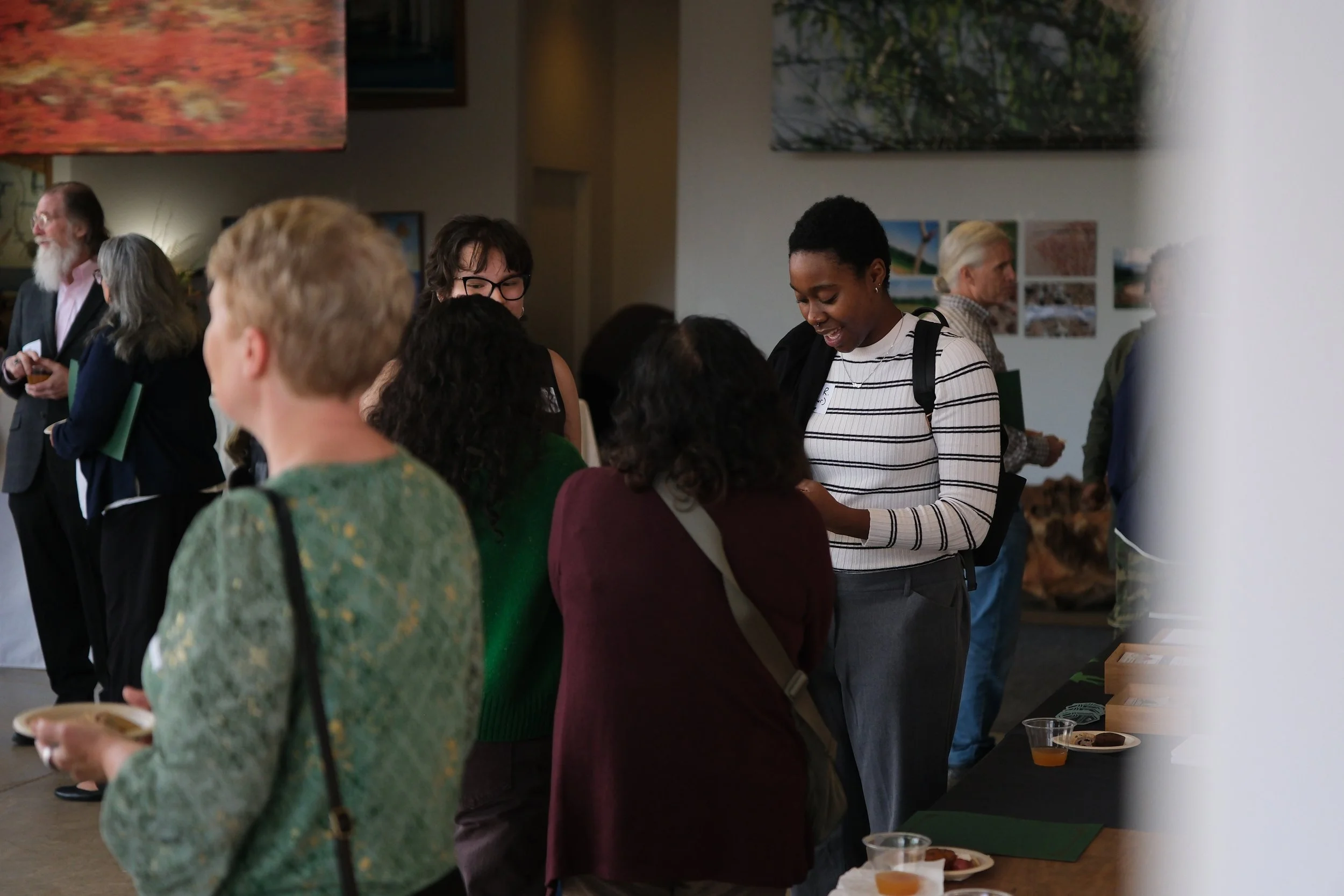 People are gathered at an indoor event, engaging in conversations and looking at display tables with food and informational materials. The setting appears to be a community or networking event with artwork on the walls.
