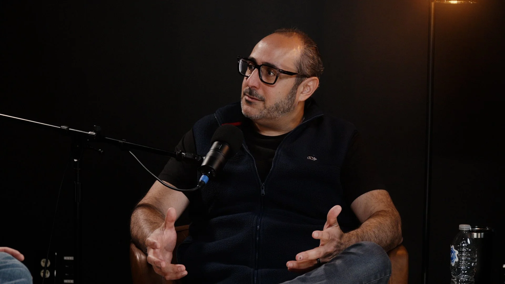A man with glasses and a beard, wearing a black vest over a black shirt, is seated in front of a black background. He is speaking into a microphone and gesturing with his hands.