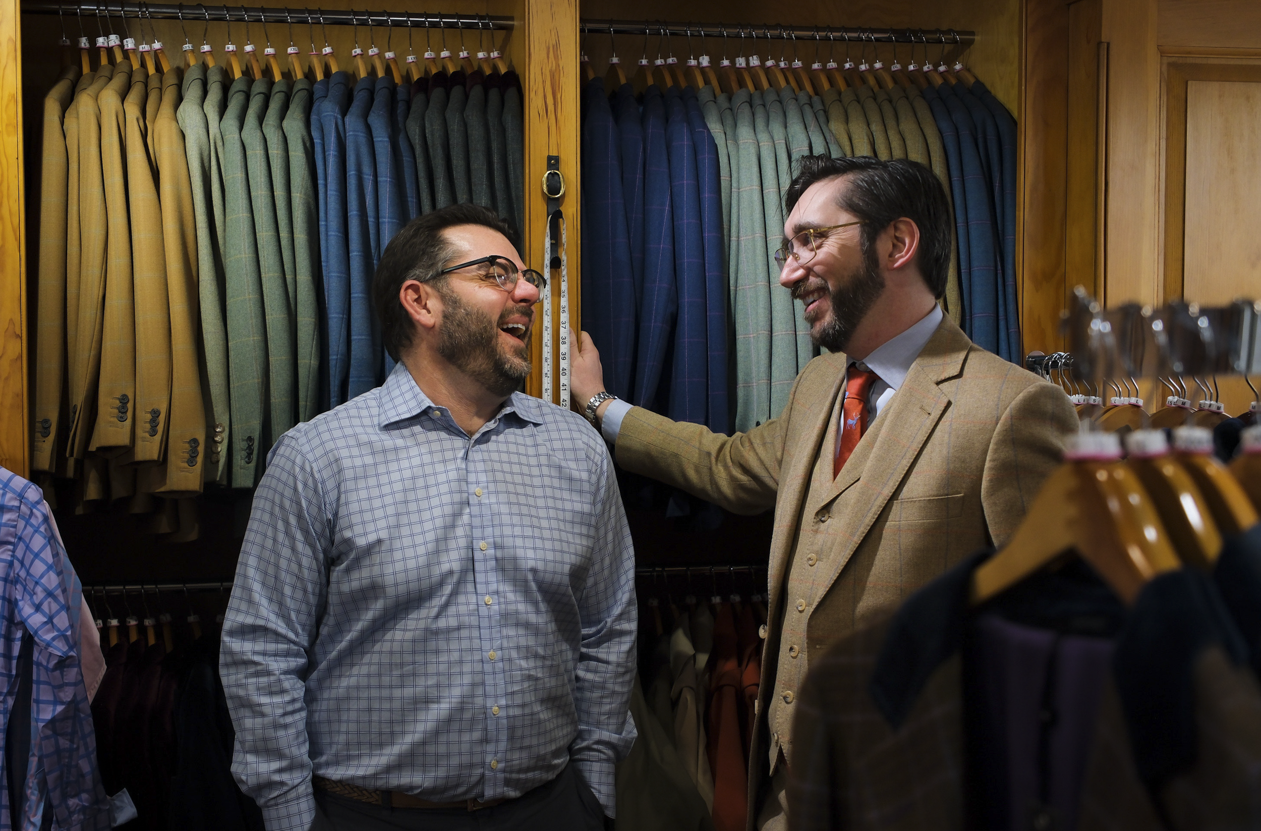 Two men with glasses and beards smiling and talking in a clothing store surrounded by racks of suits and jackets.
