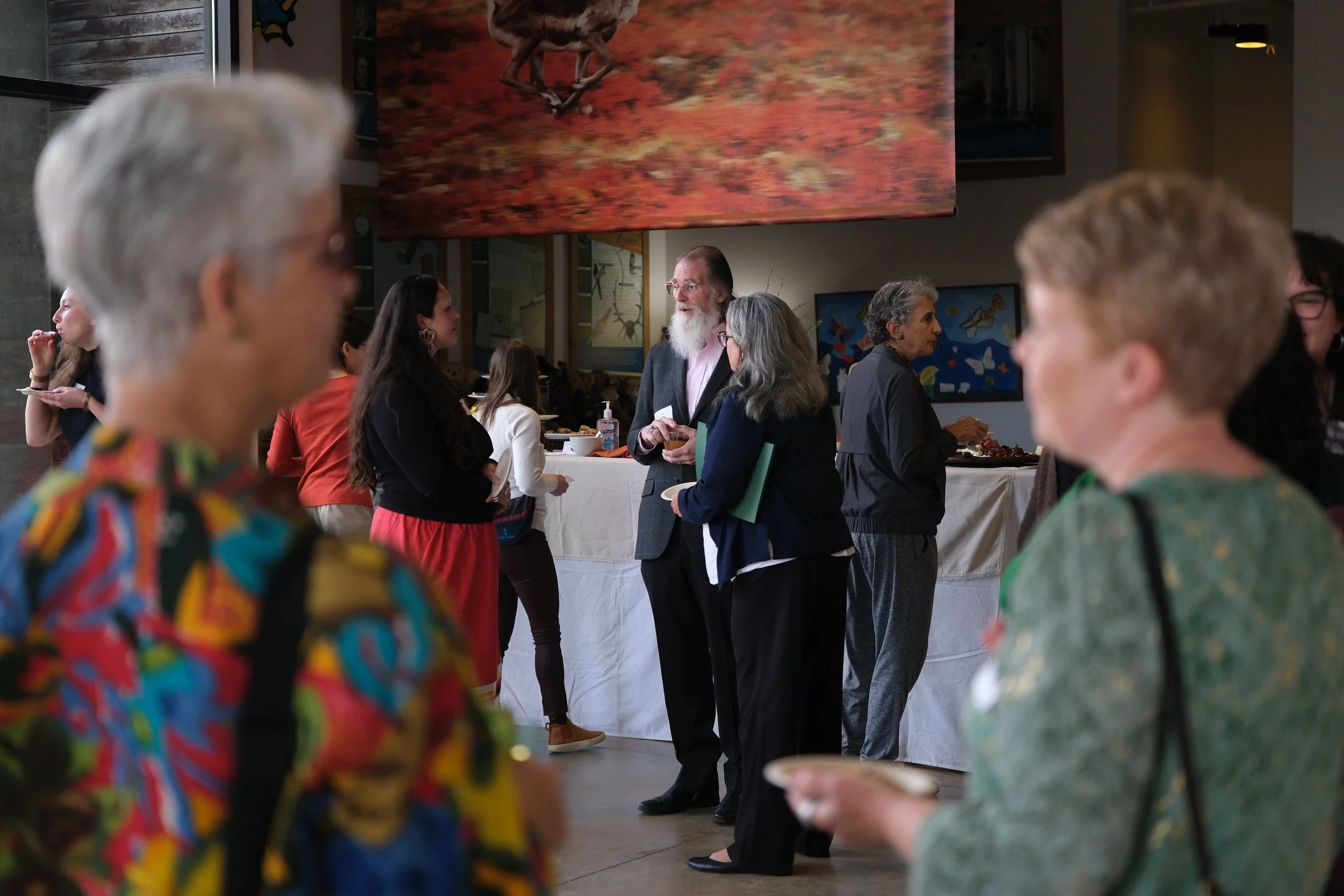 People gathered at an indoor event, engaging in conversations near a buffet table, with colorful artwork and a large painting on the wall in the background.